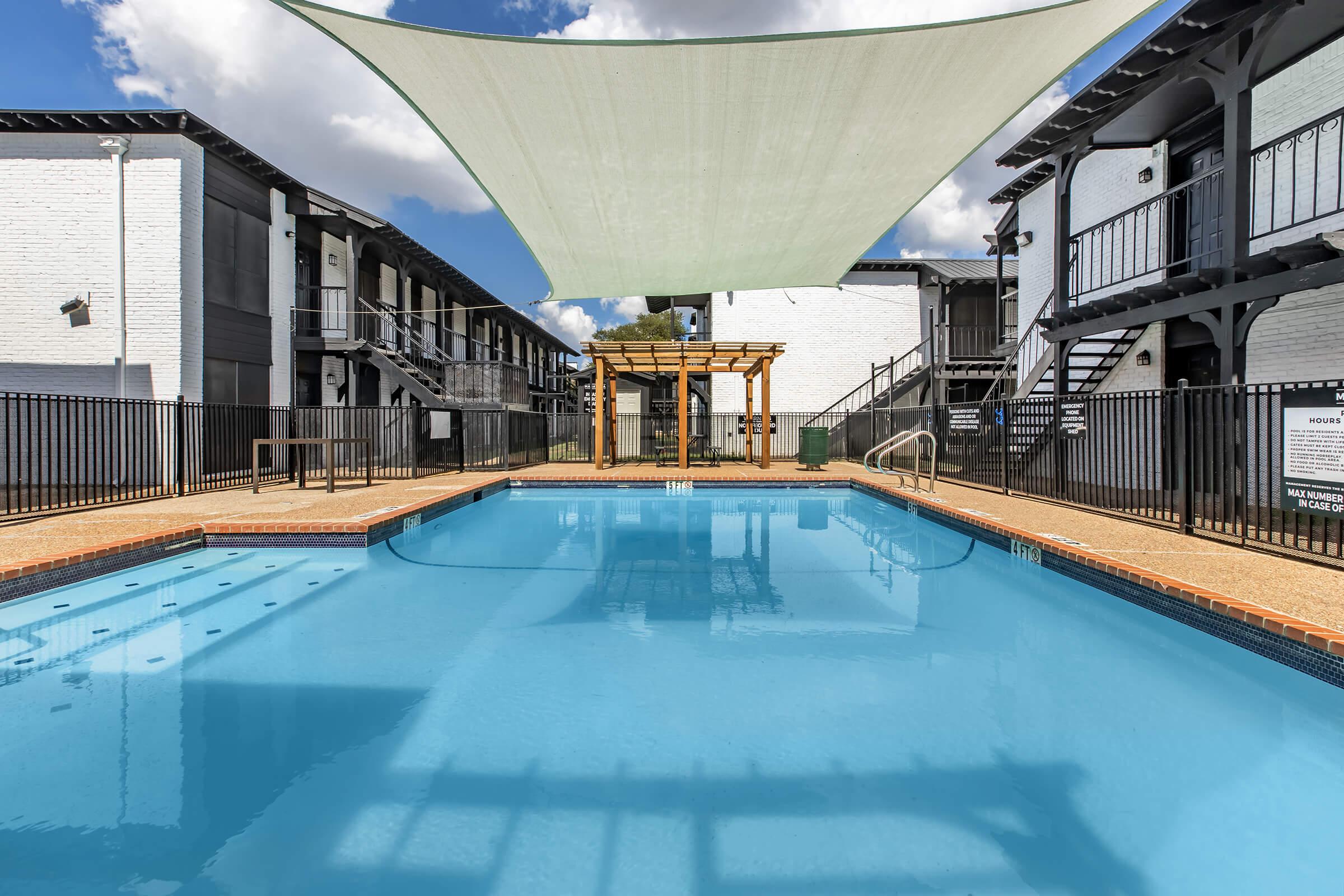 A clear blue swimming pool surrounded by brick decking, with a shaded area provided by a large canopy. In the background, white apartment buildings and wooden structures are visible. The scene is bright and inviting, perfect for relaxation.