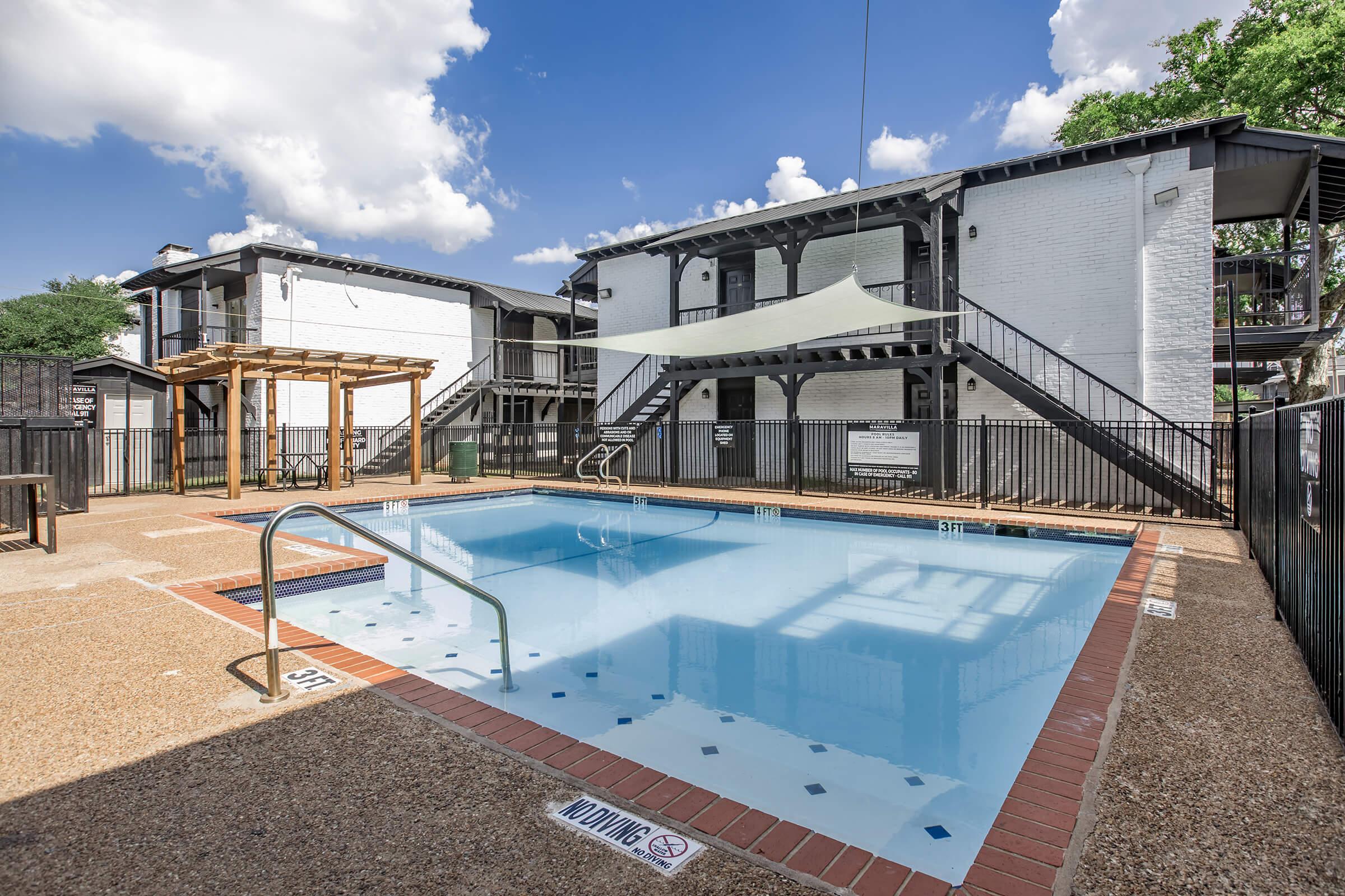 A clear swimming pool surrounded by a patio area, with a shaded seating space nearby. In the background, there are two multi-story buildings. The sky is partly cloudy, and the setting appears inviting and well-maintained, suitable for relaxation and leisure activities.
