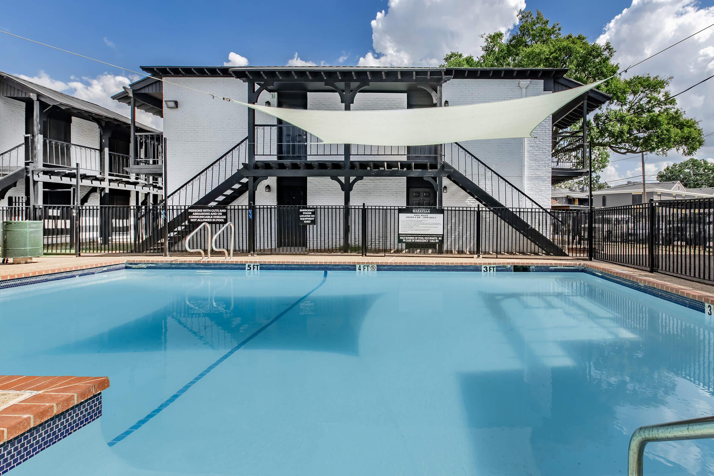 A swimming pool surrounded by a black fence, with a sunshade overhead. In the background, there is a two-story white building with stairs leading to the upper level. The pool is clear and inviting, reflecting the blue sky and clouds above.