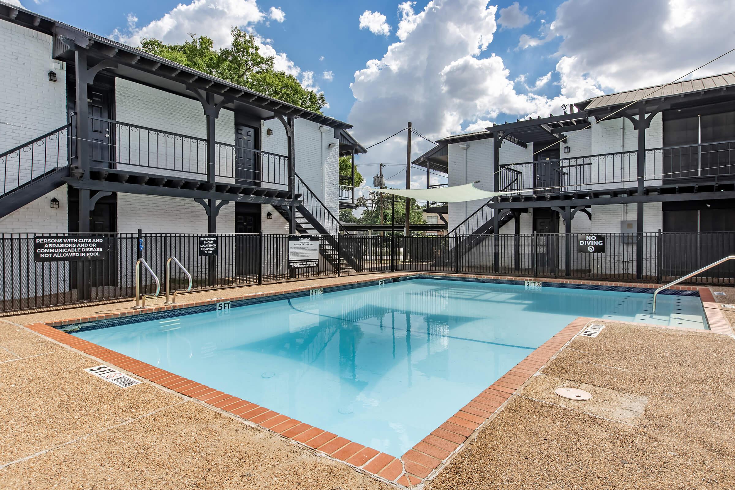 A rectangular swimming pool surrounded by a gated area, set between two white, two-story apartment buildings. Lush greenery in the background and scattered clouds in a blue sky. The pool area includes steps leading into the water and signs indicating pool rules.
