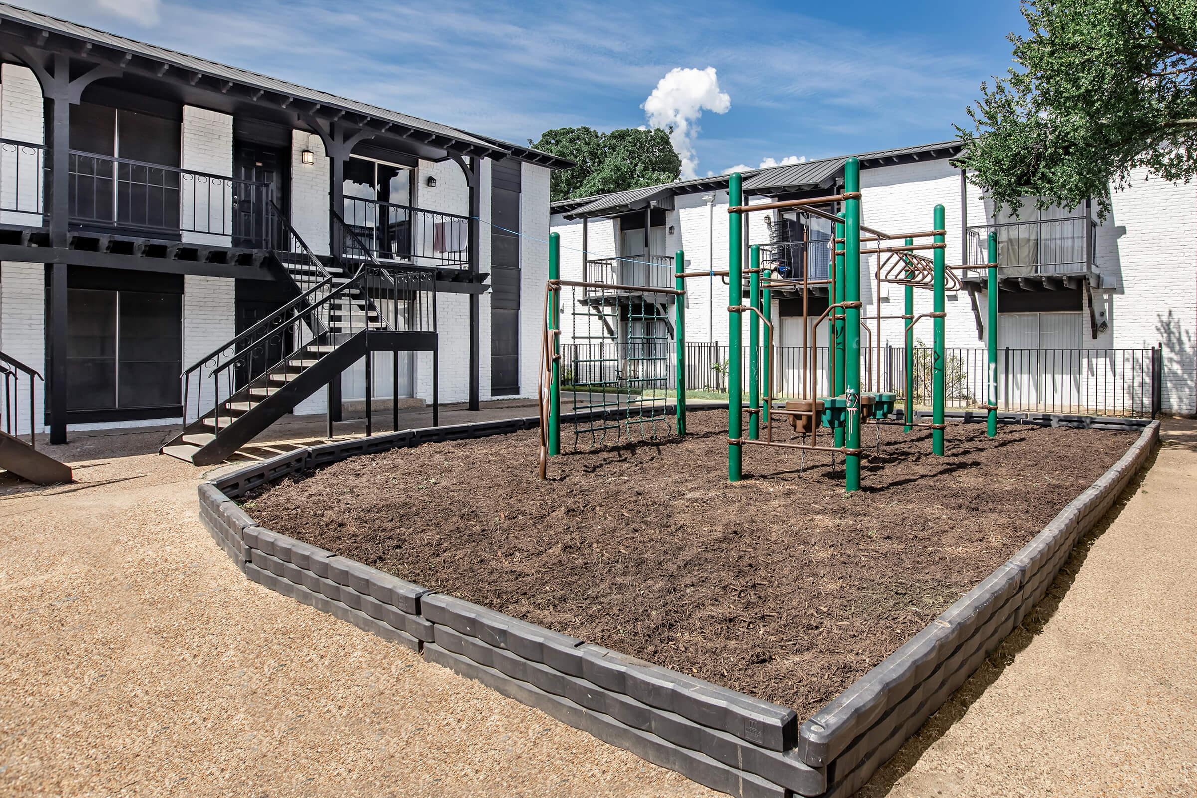 A playground area featuring green climbing structures surrounded by a gravel path and bordered by raised planters. The background includes a two-story apartment building with a wooden staircase and balcony access. Clear blue skies and a few clouds are visible above.