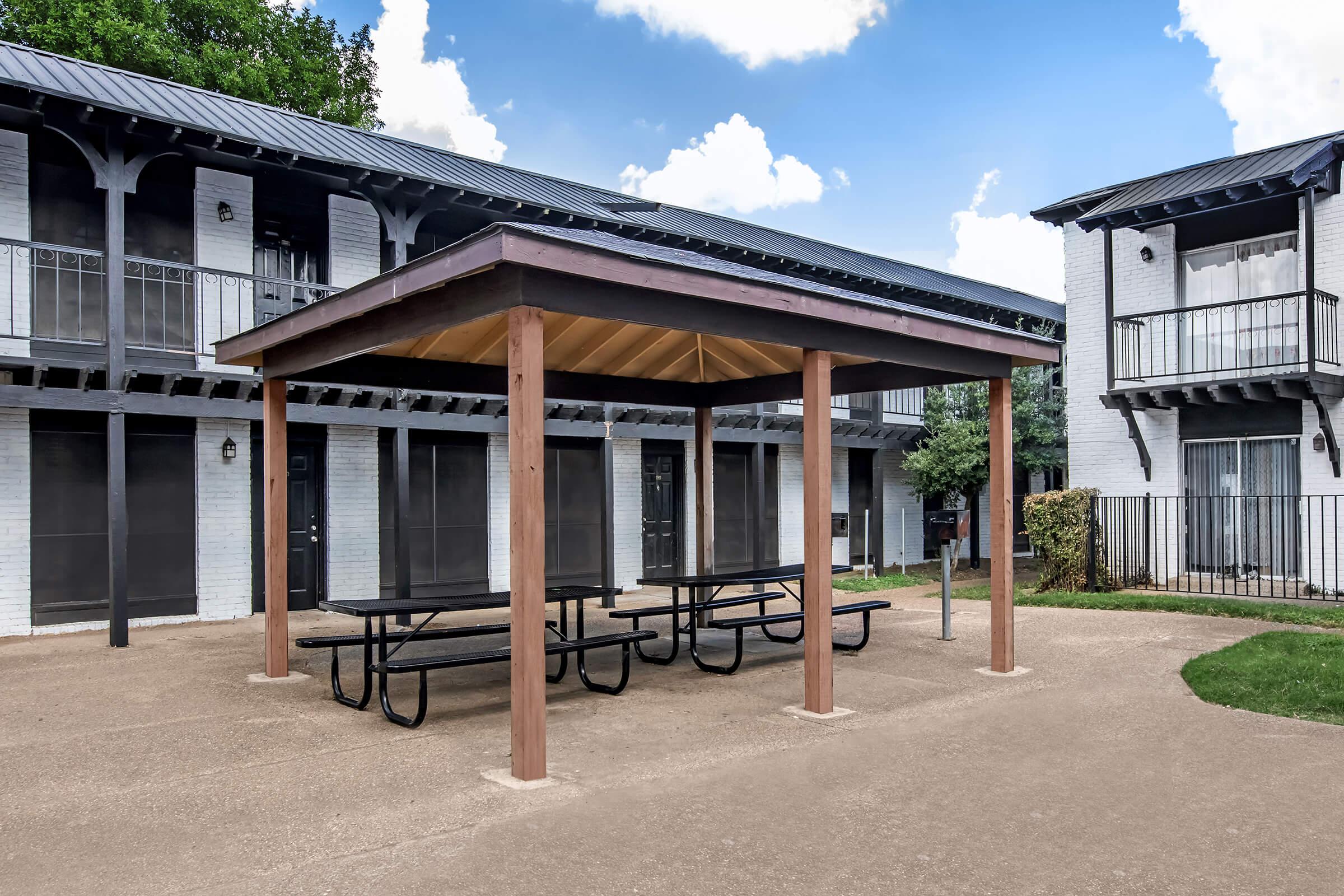 A covered picnic area with wooden beams and a metal roof is situated between two multi-story residential buildings. Two picnic tables are placed underneath the shelter, and the surrounding pavement is well-maintained, with grass and shrubs in the background under a partly cloudy sky.