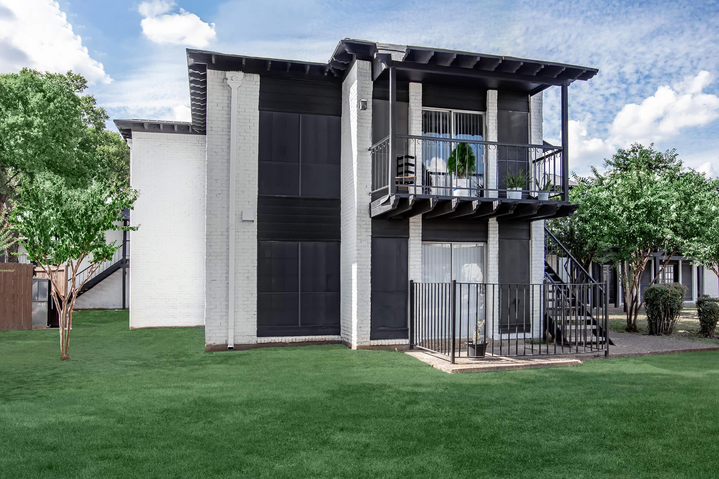 A modern two-story apartment building with a black and white facade. The upper floor features a balcony, while the lower floor has a gated patio area. Surrounding the building is a well-manicured lawn with small trees, set against a blue sky with fluffy clouds.