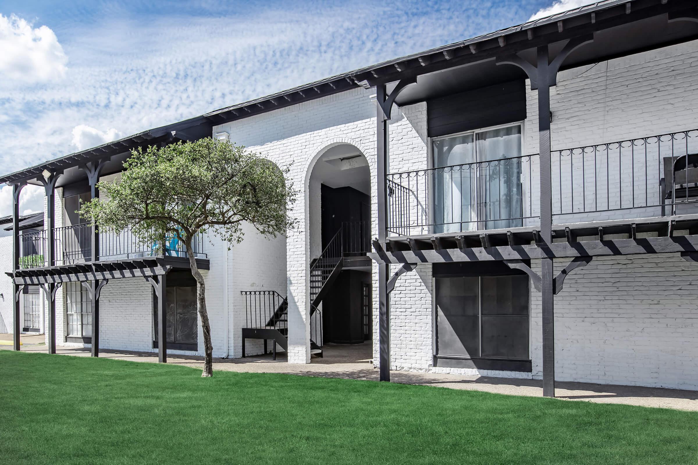A modern two-story building with white brick walls and black wooden accents. The structure features arched doorways and balconies with railings. A small tree is placed in the foreground on a manicured green lawn under a bright blue sky with some clouds.