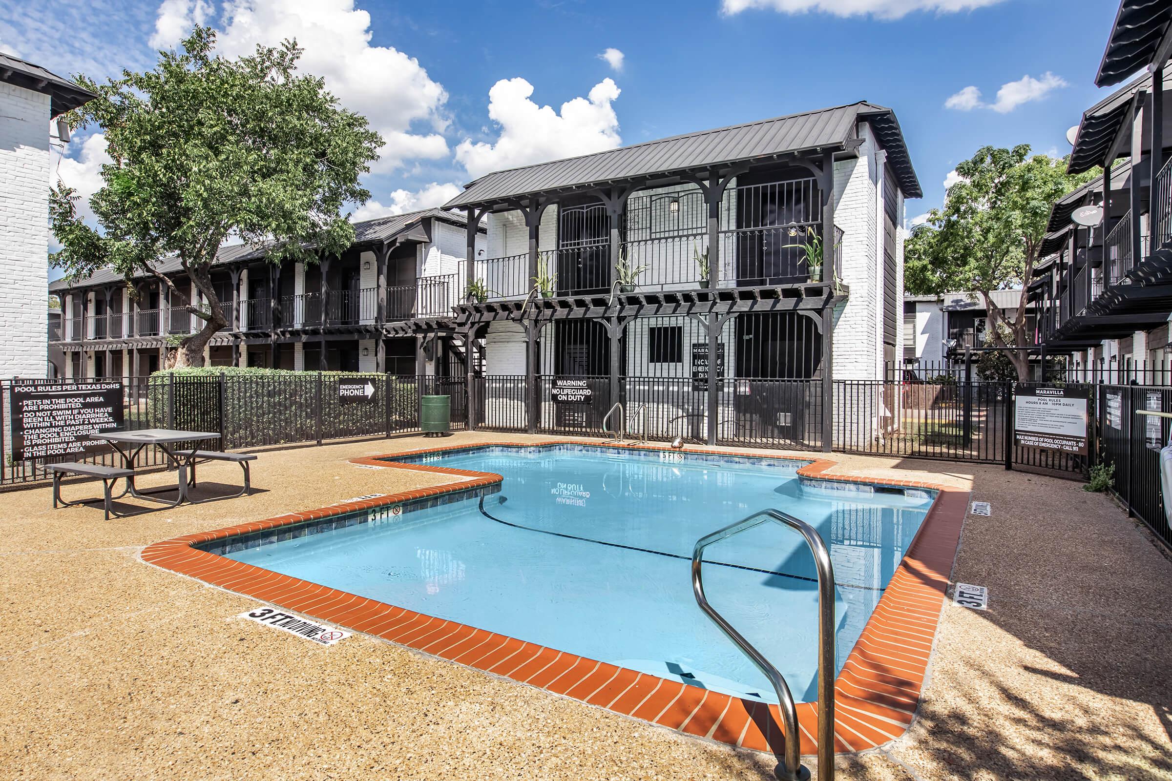 A serene swimming pool surrounded by a fenced area, with lounge chairs positioned nearby. The backdrop features a two-story apartment building with balconies and lush greenery under a bright blue sky dotted with fluffy clouds.
