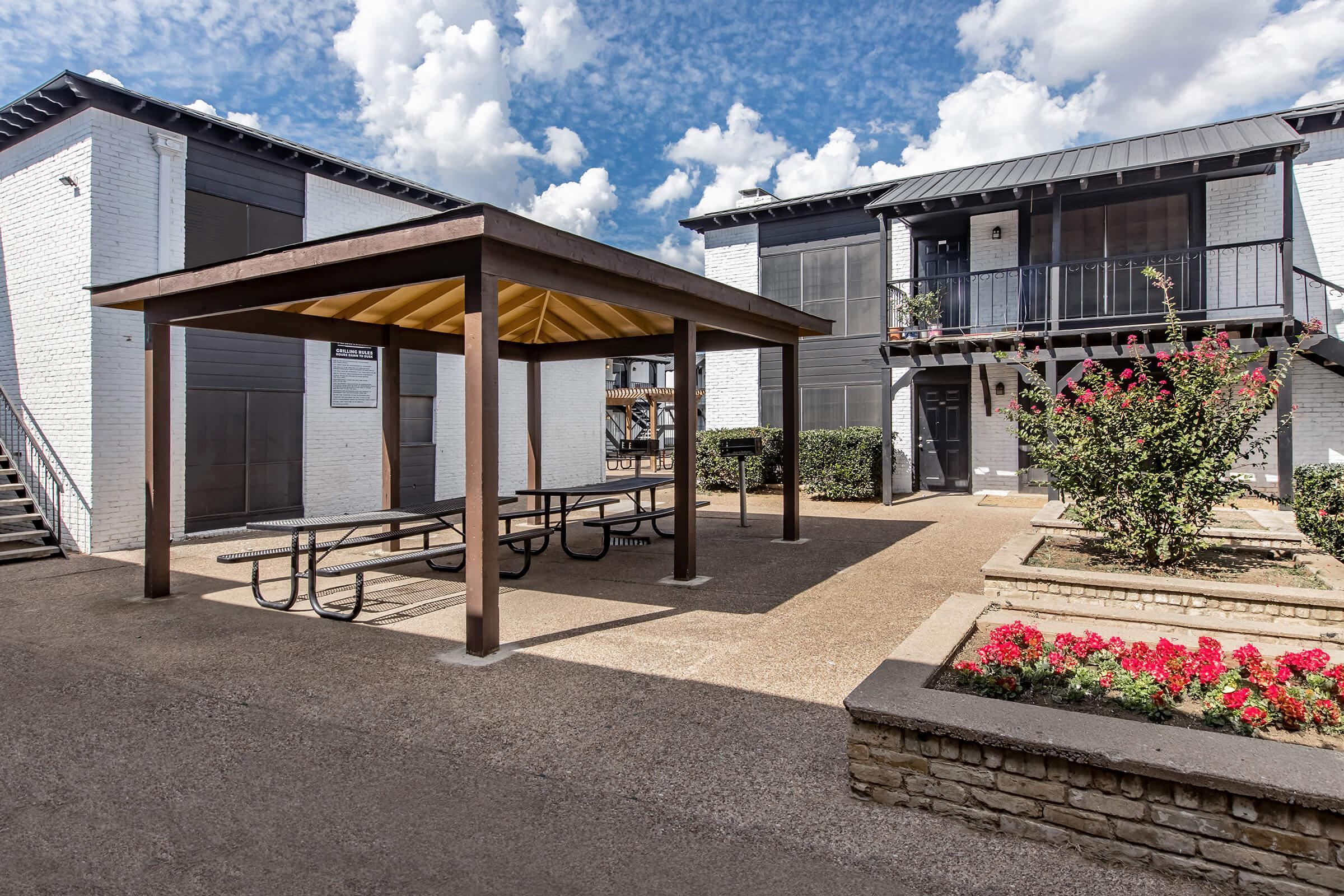 A courtyard scene featuring a two-story apartment building with a modern design, a covered picnic area with tables, and landscaped flower beds with colorful blooms. The sky is partly cloudy, creating a bright atmosphere. The walkway is paved and leads to the entrance of the building.