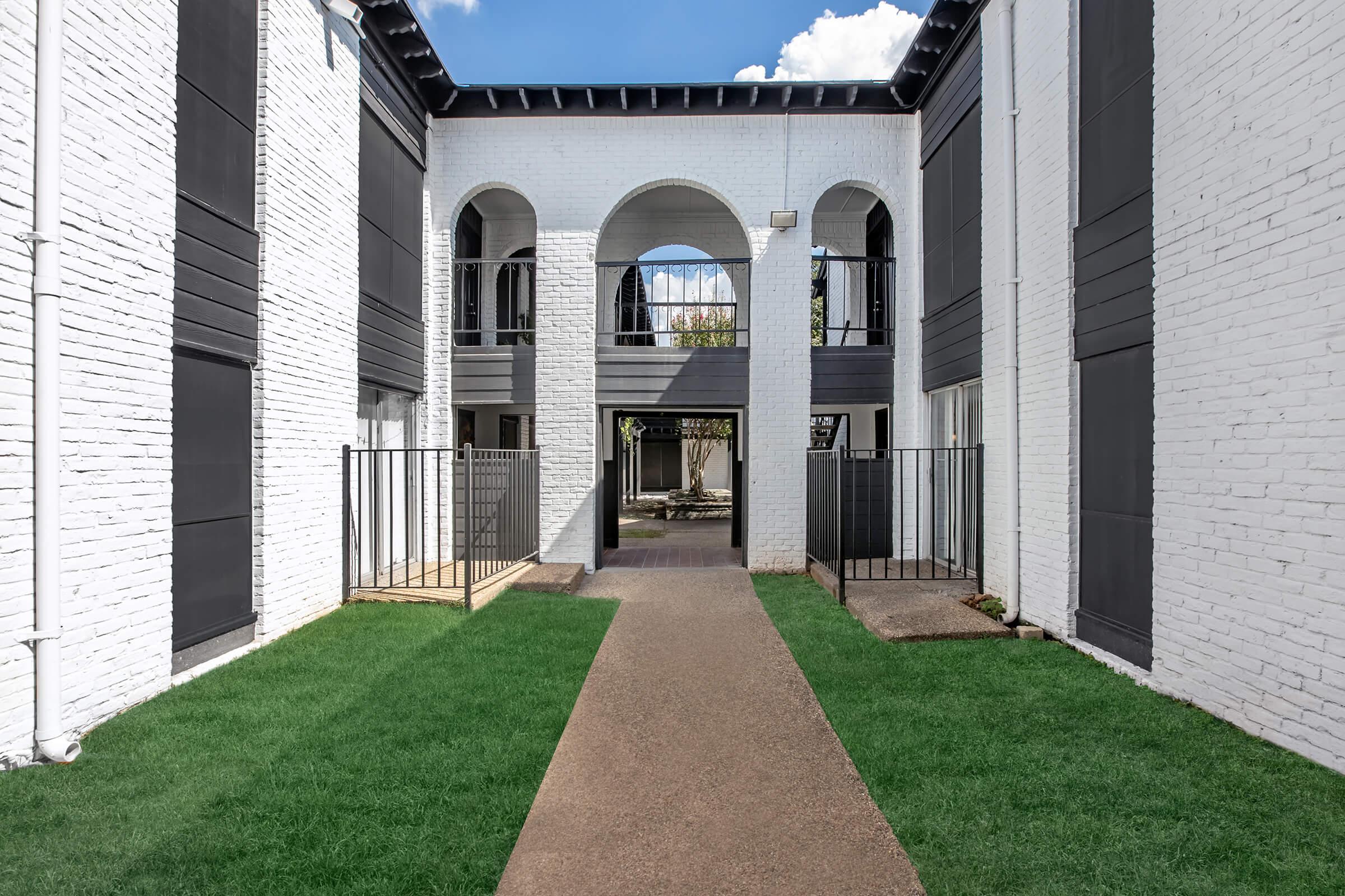 A well-maintained outdoor pathway lined with green grass, leading through an archway between two white brick buildings. The architecture features black accents and balconies. The scene is bright, with blue skies and fluffy clouds visible above.