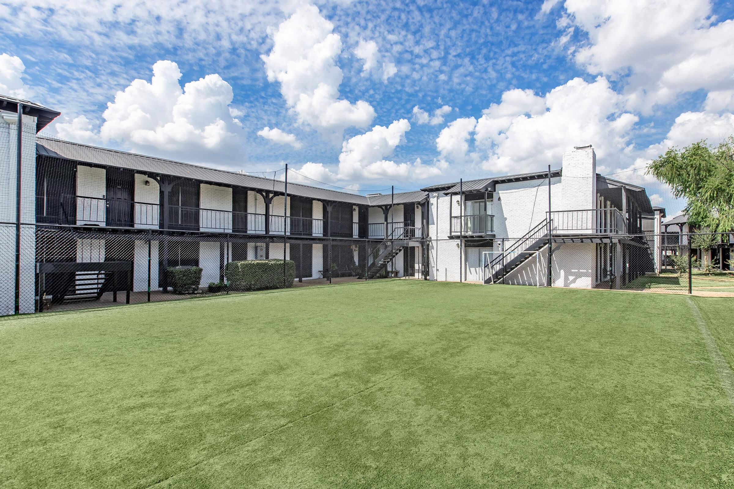 View of a two-story building with balconies facing a green lawn, surrounded by a chain-link fence. The sky is bright and partly cloudy, creating a vibrant atmosphere. The setting appears to be a well-maintained outdoor space, suitable for recreational activities.