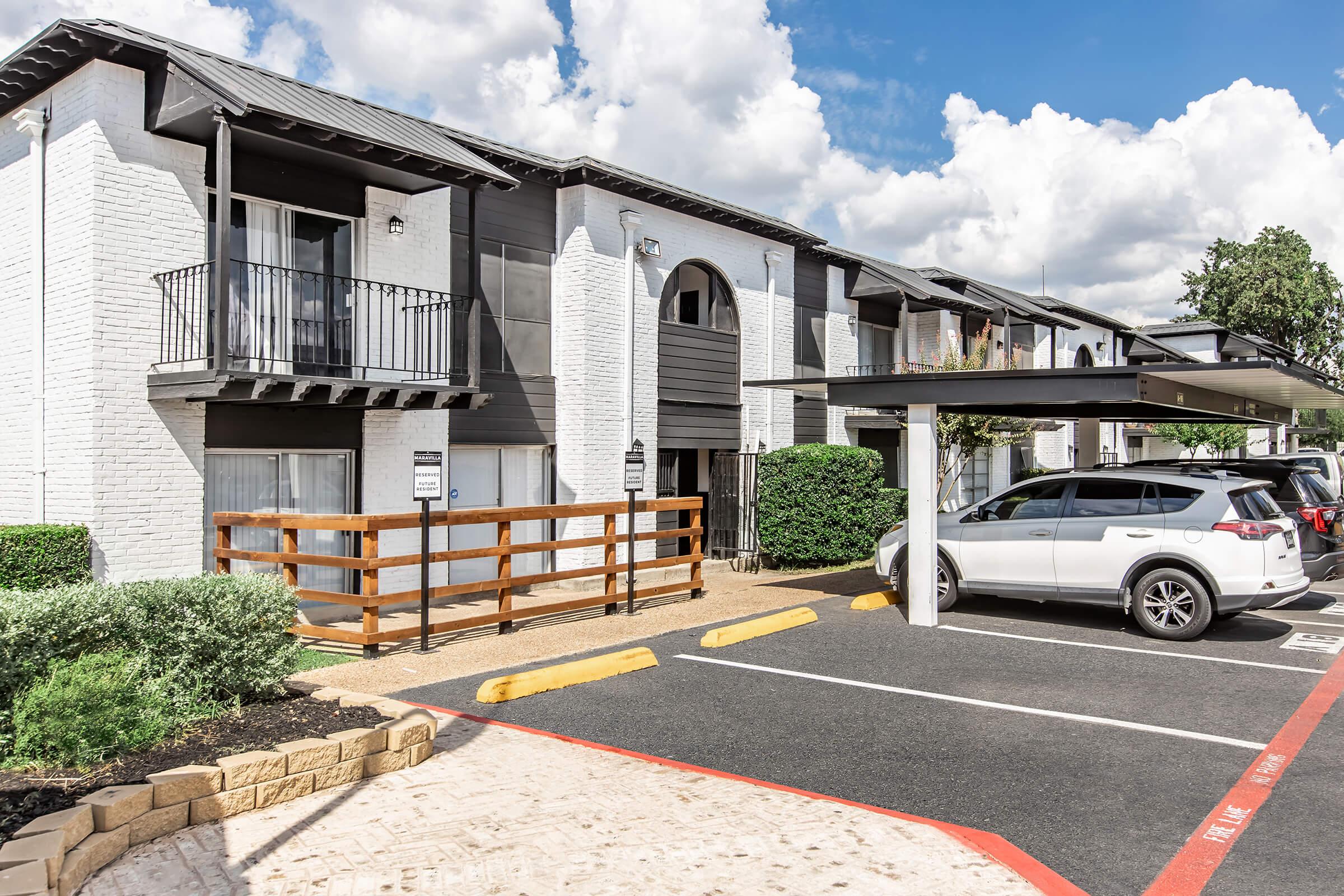 A two-story apartment building with a modern design featuring white brick and dark accents. There are balconies on the second floor, a wooden fence in front, and a covered parking area. Several cars are parked nearby, and the background includes blue skies with fluffy clouds.