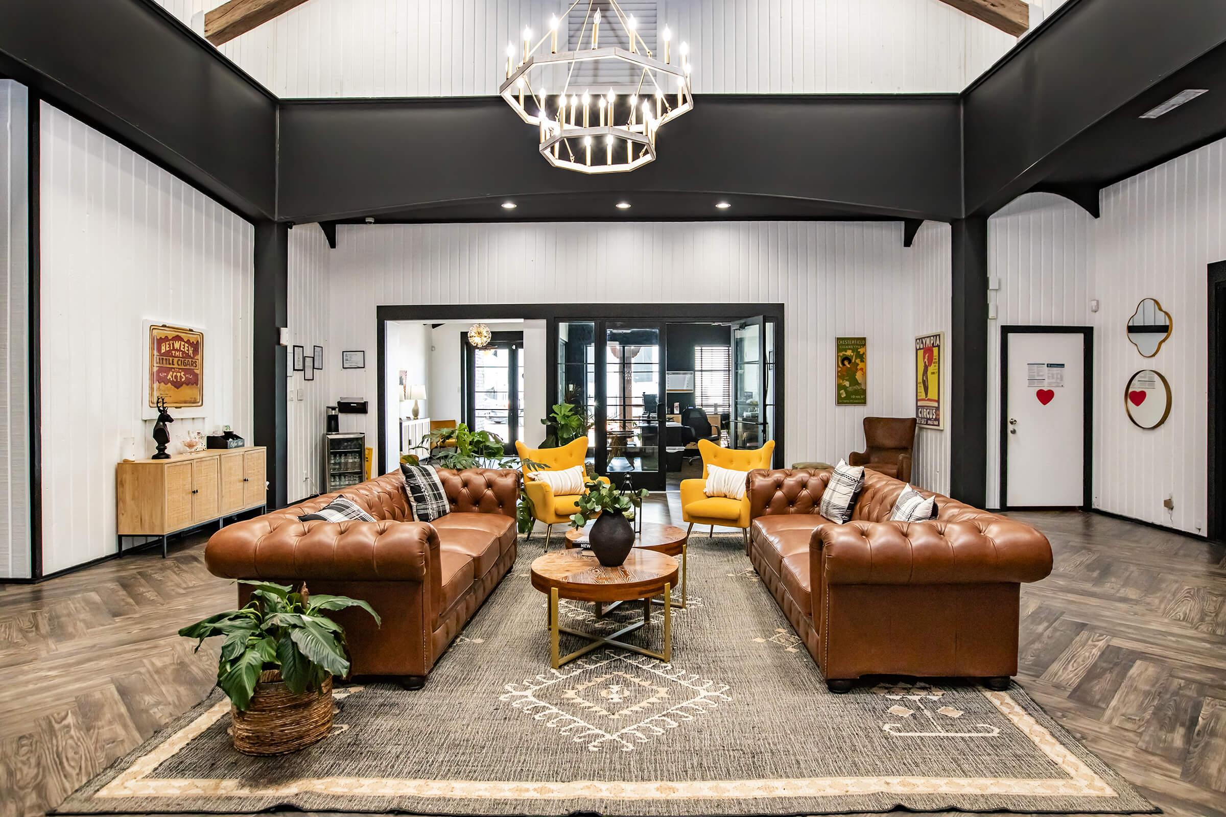 A spacious living area featuring two brown leather chesterfield sofas, a round coffee table, and a patterned rug. Large windows allow natural light, while potted plants and vibrant yellow chairs add warmth. Walls are adorned with artwork, and a modern chandelier hangs from the high ceiling.