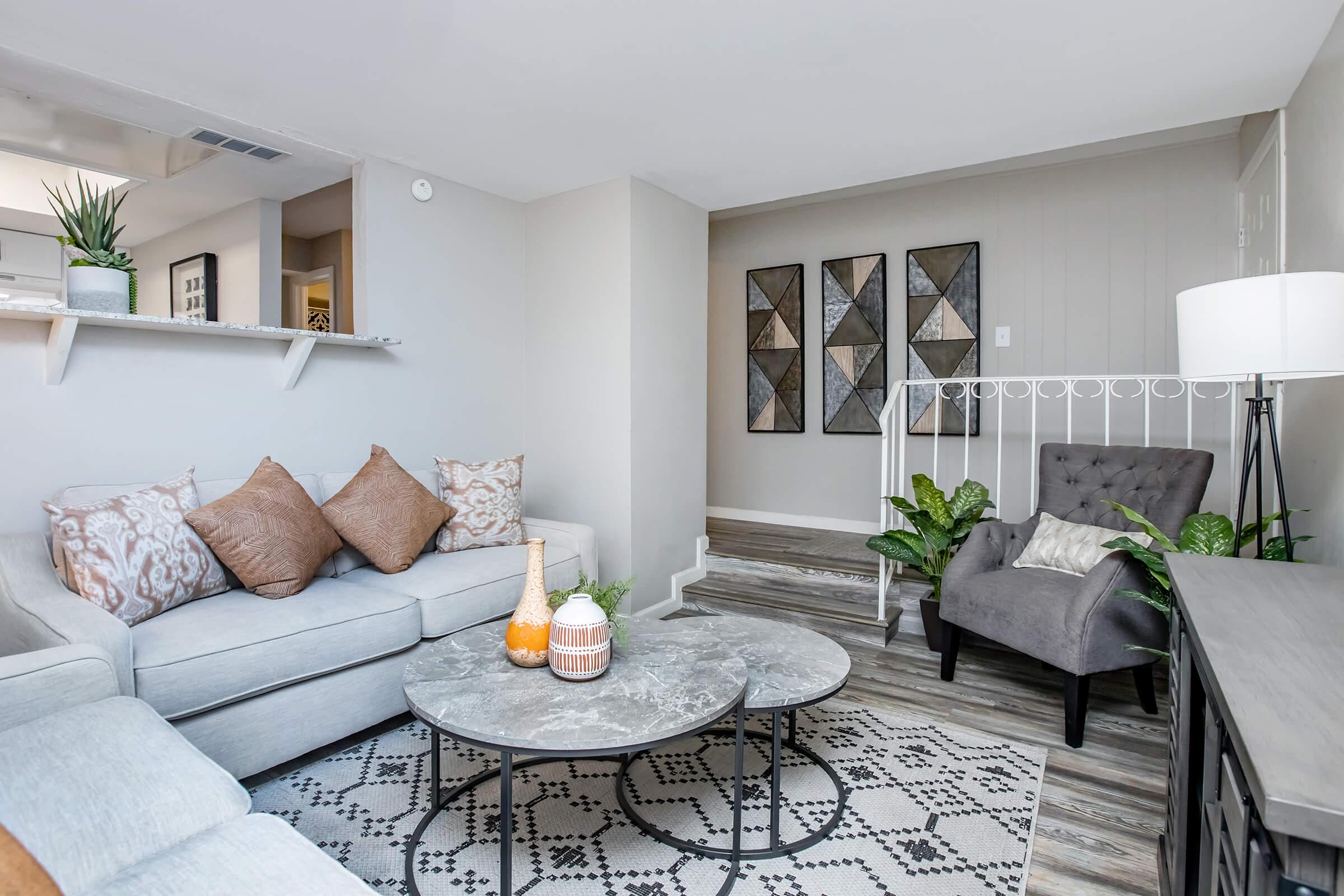 A modern living room featuring a light gray sofa with decorative pillows, a round marble coffee table, and a gray armchair. A patterned area rug covers the floor, and the walls are light-colored with geometric wall art. Potted plants add greenery, and a lamp stands beside the armchair. Stairs lead to another level.