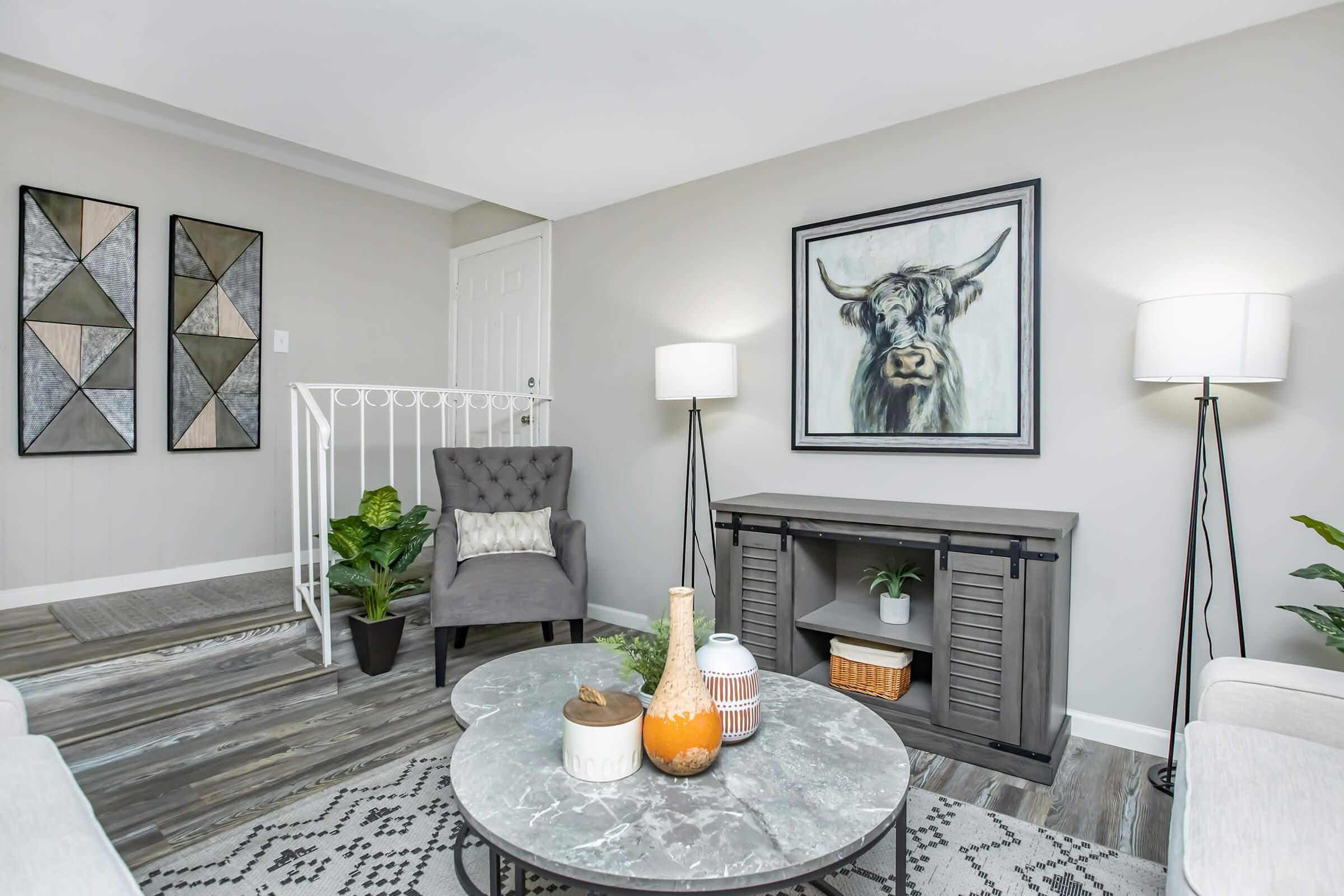 A modern living room featuring a gray armchair, a coffee table with decorative items, and a stylish console table. The walls are light gray, adorned with a cow portrait and geometric artwork. Two floor lamps provide soft lighting, while a plant adds a touch of greenery to the space.