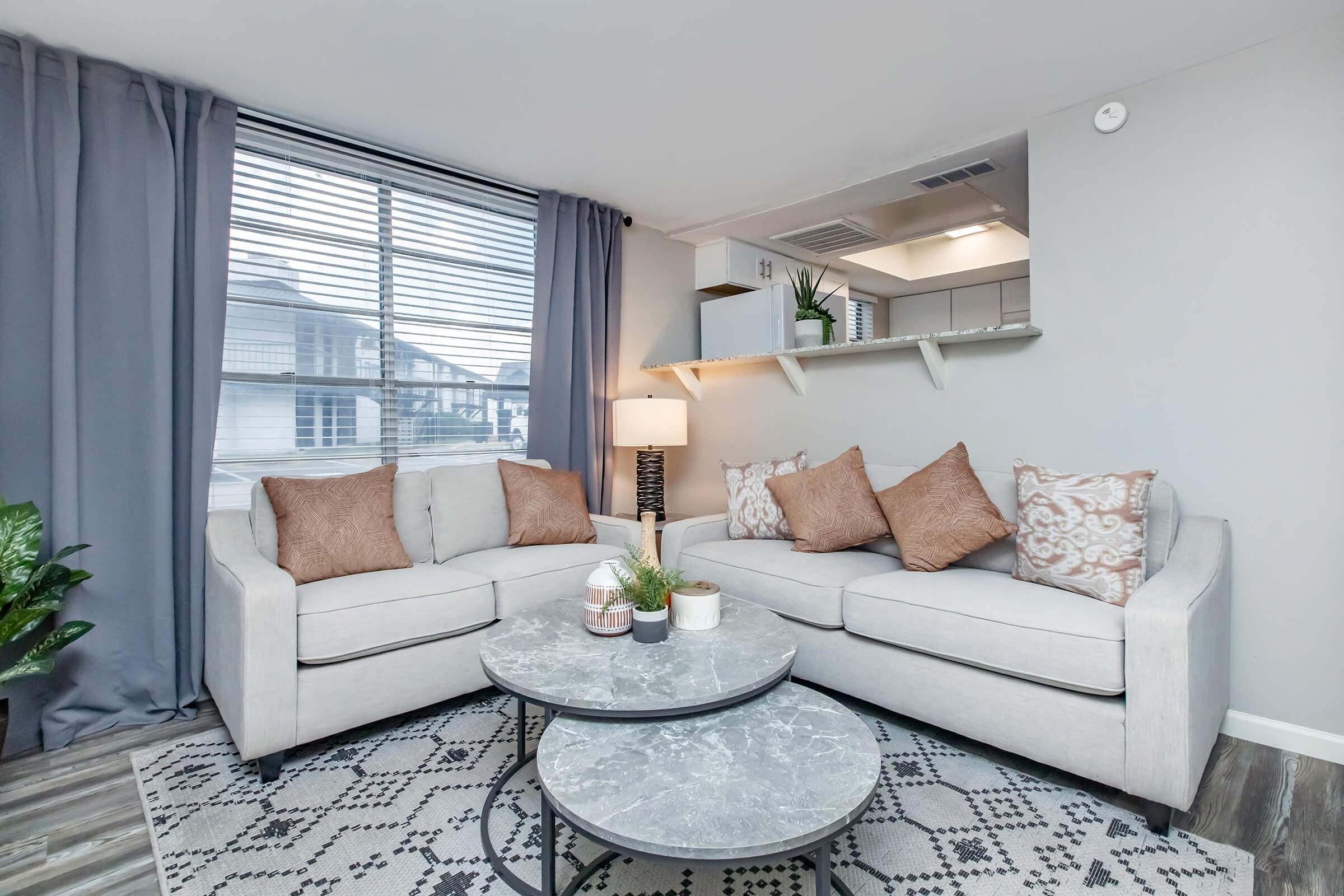 A cozy living room featuring a light gray couch adorned with decorative pillows, a round marble coffee table, and a potted plant. Natural light streams through a window with sheer curtains. The room has a modern decor style, including soft lighting and a shelf with plants in the background.