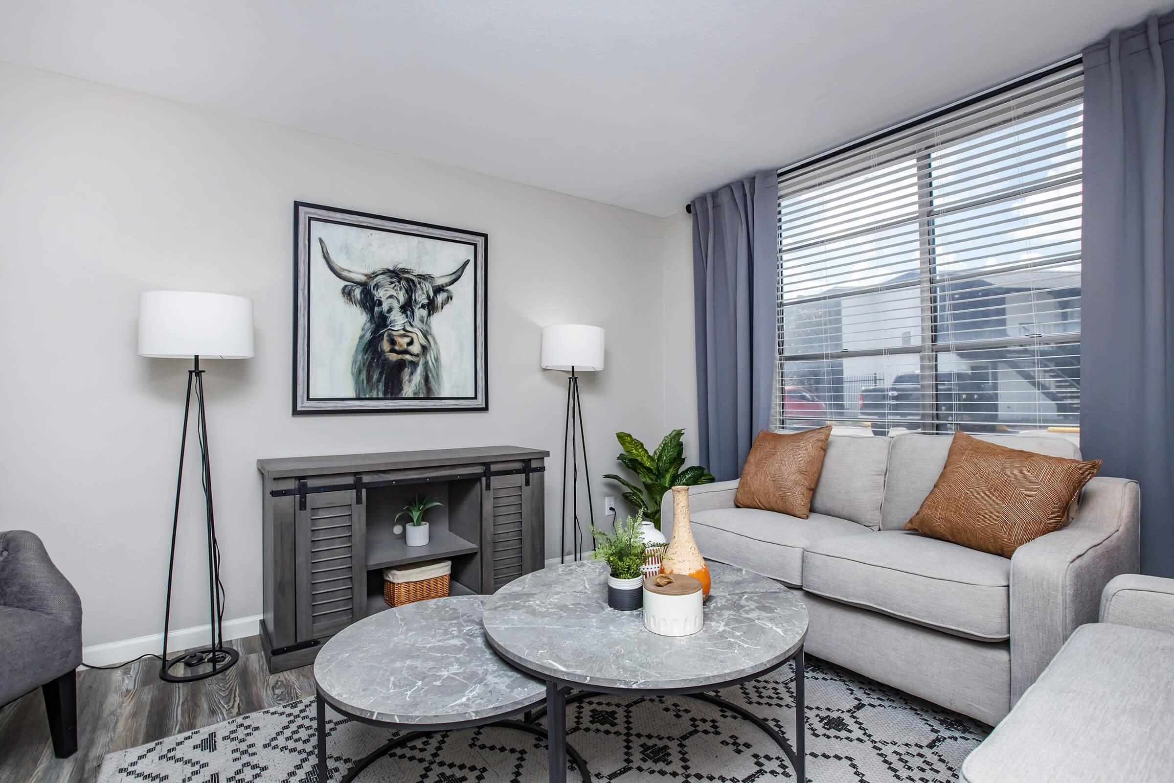 Cozy living room featuring a gray sofa with decorative pillows, a coffee table with a marble top, and two stylish floor lamps. A large framed painting of a cow hangs on the wall. The room is illuminated by natural light from a window covered with sheer curtains, adding warmth to the modern decor.
