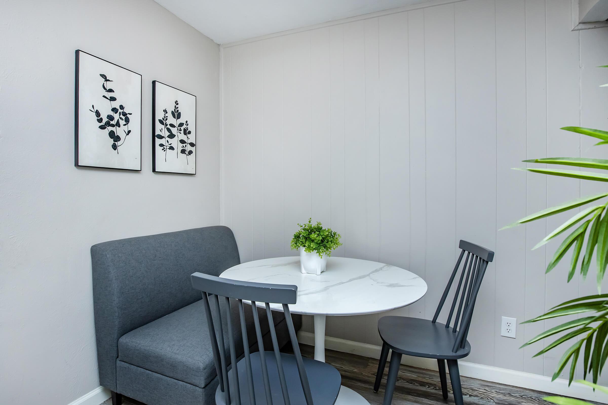 A cozy dining area featuring a round marble table surrounded by two black chairs and a gray bench. The walls are painted light gray and adorned with two framed botanical prints. A small potted plant sits on the table, adding a touch of greenery to the space.
