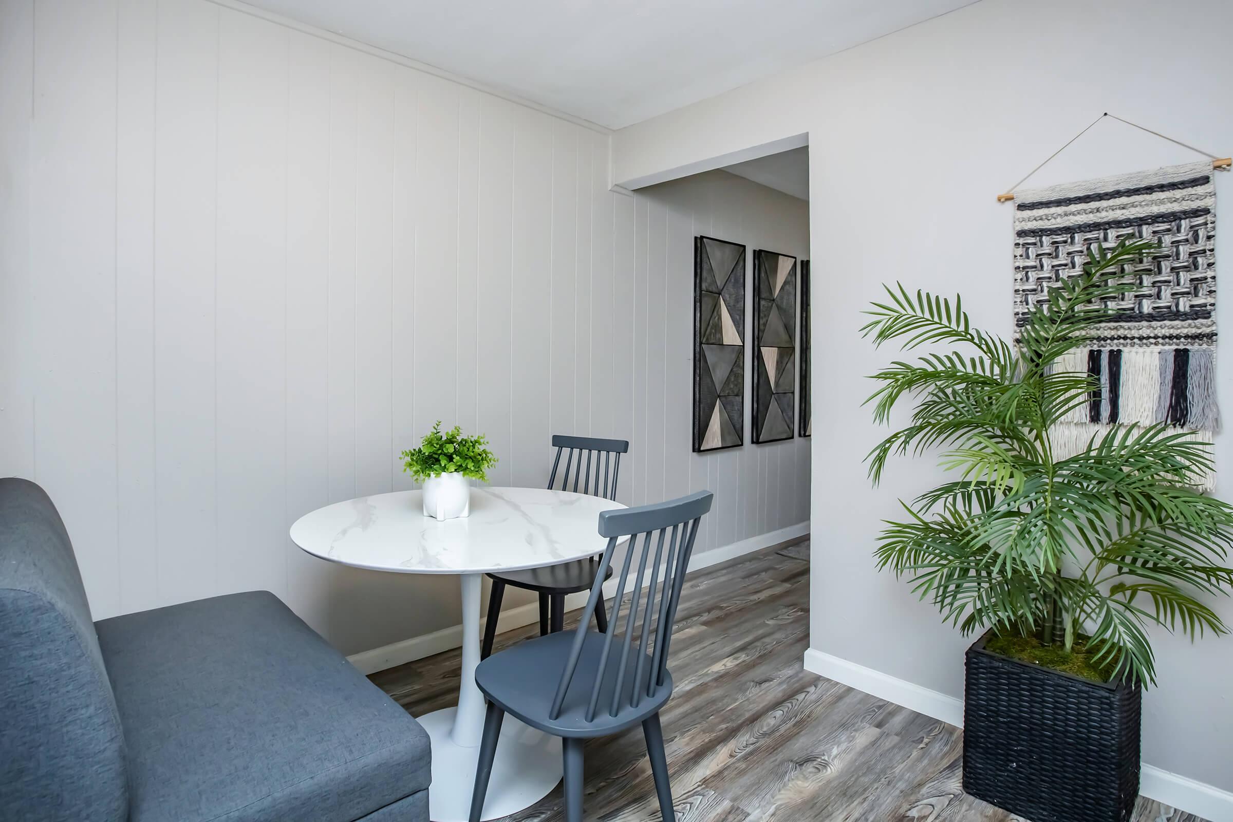 A cozy dining area featuring a round marble table with two gray chairs, a small potted plant on the table, and a gray upholstered bench. The walls are light-colored with decorative wall art and a woven wall hanging. A large green plant in a black pot adds a touch of nature to the space.