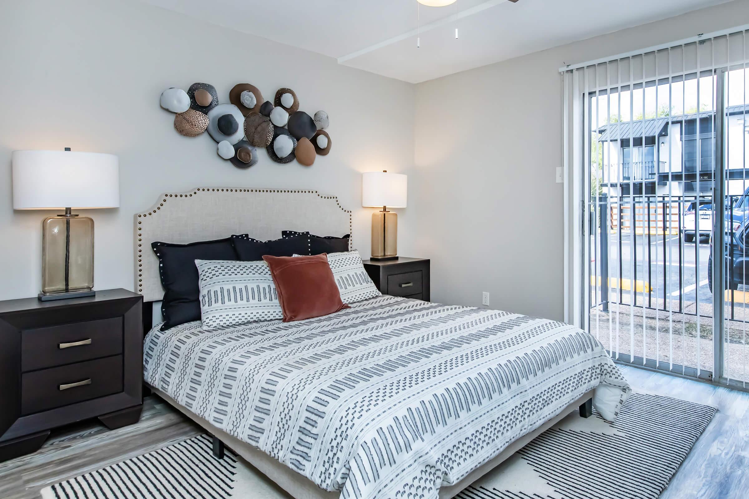 A cozy bedroom featuring a bed with a patterned duvet and a decorative pillow. There are two stylish bedside tables with lamps, and wall art made of circular shapes above the bed. A window with vertical blinds allows natural light, and there’s visible outdoor space through the window.