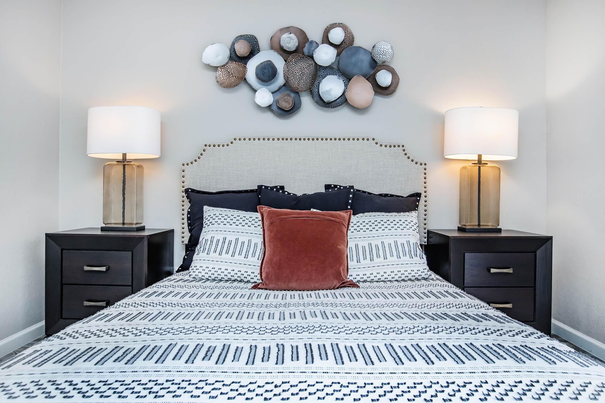 A stylish bedroom featuring a neatly made bed with patterned bedding and decorative pillows. On either side of the bed are dark wooden nightstands with lamps. Above the headboard hangs a collection of decorative wall art made of various shapes and textures. The overall color scheme is neutral with pops of color.