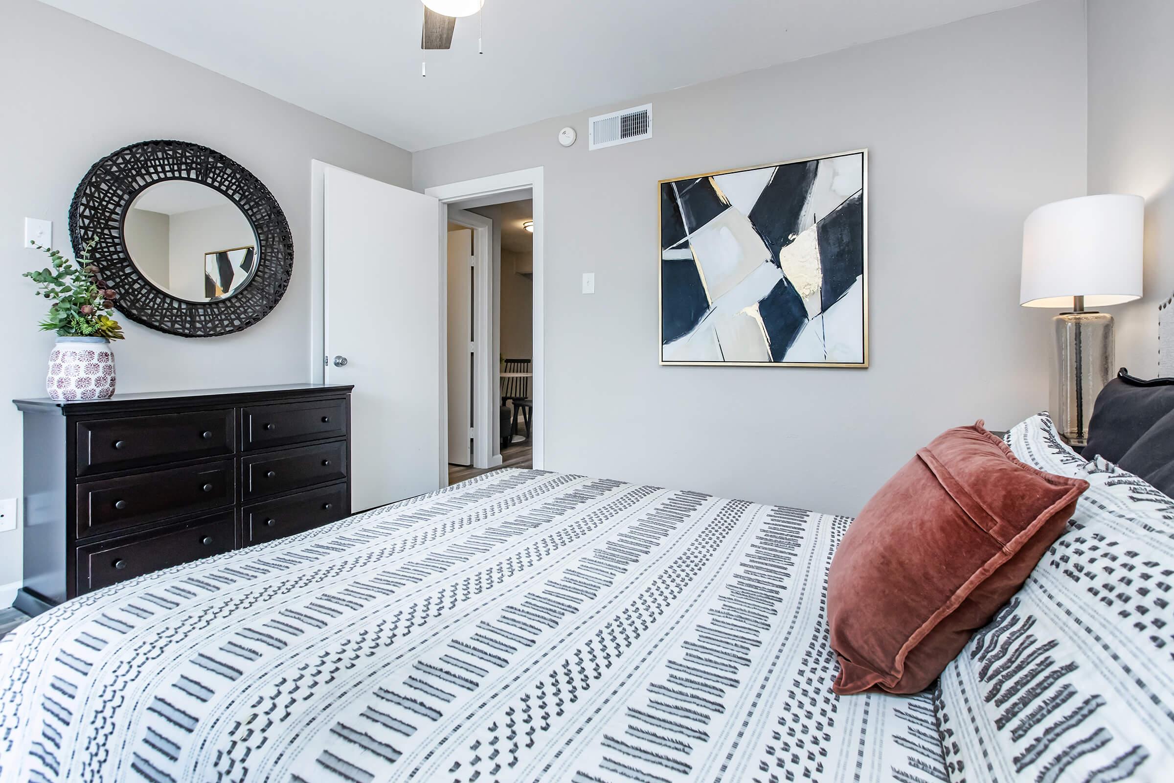 A well-decorated bedroom featuring a bed with a patterned blanket and decorative pillows. There's a round mirror on the wall, a stylish piece of abstract art, a modern lamp on a nightstand, and a dresser. The walls are a soft gray, giving the room a cozy and inviting ambiance.
