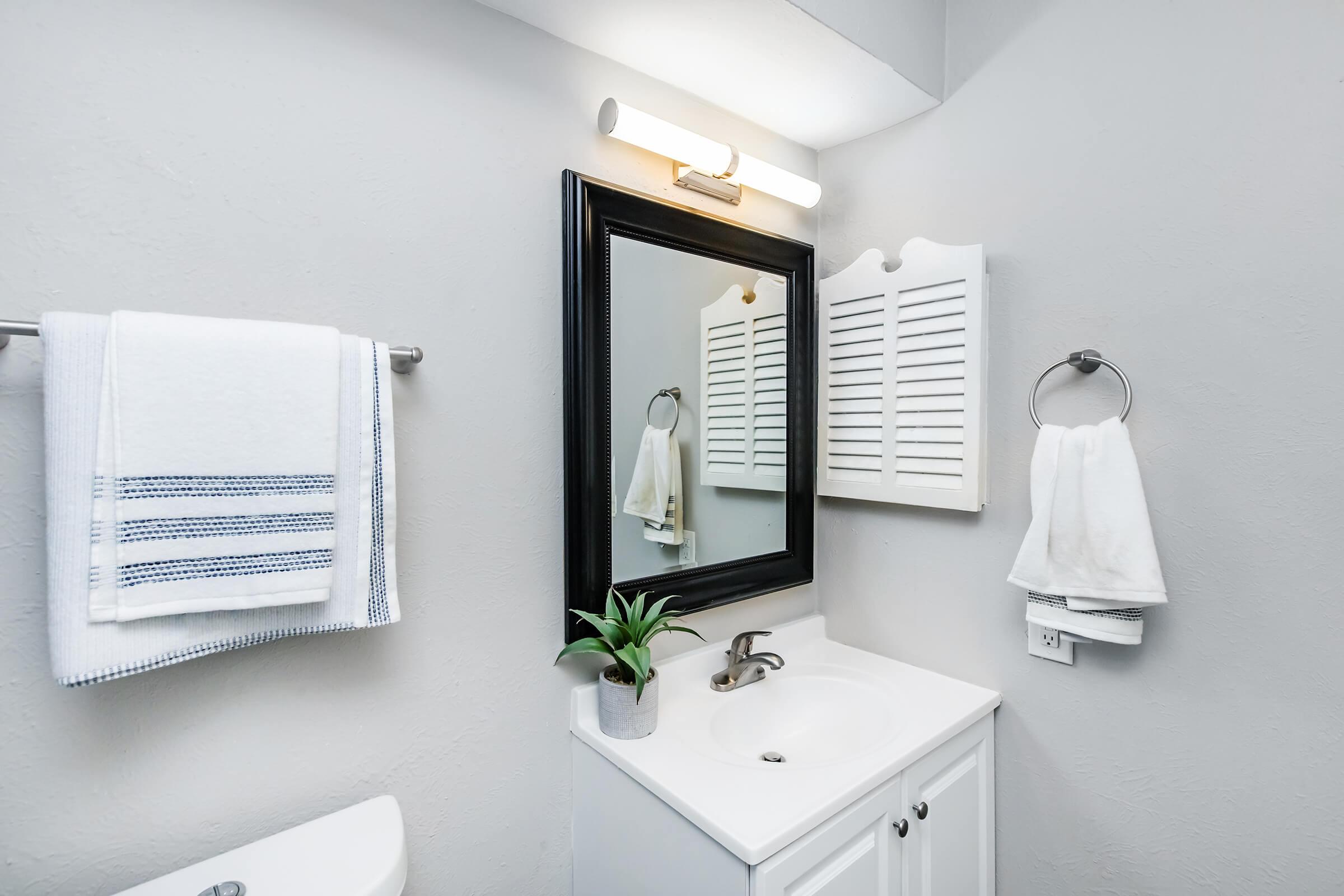 A modern bathroom featuring a white sink with a black-framed mirror above it. There are two white towels hanging on towel bars, a small plant on the counter, and a light fixture above the mirror. The walls are painted a light gray, and there is a decorative cabinet beside the mirror.