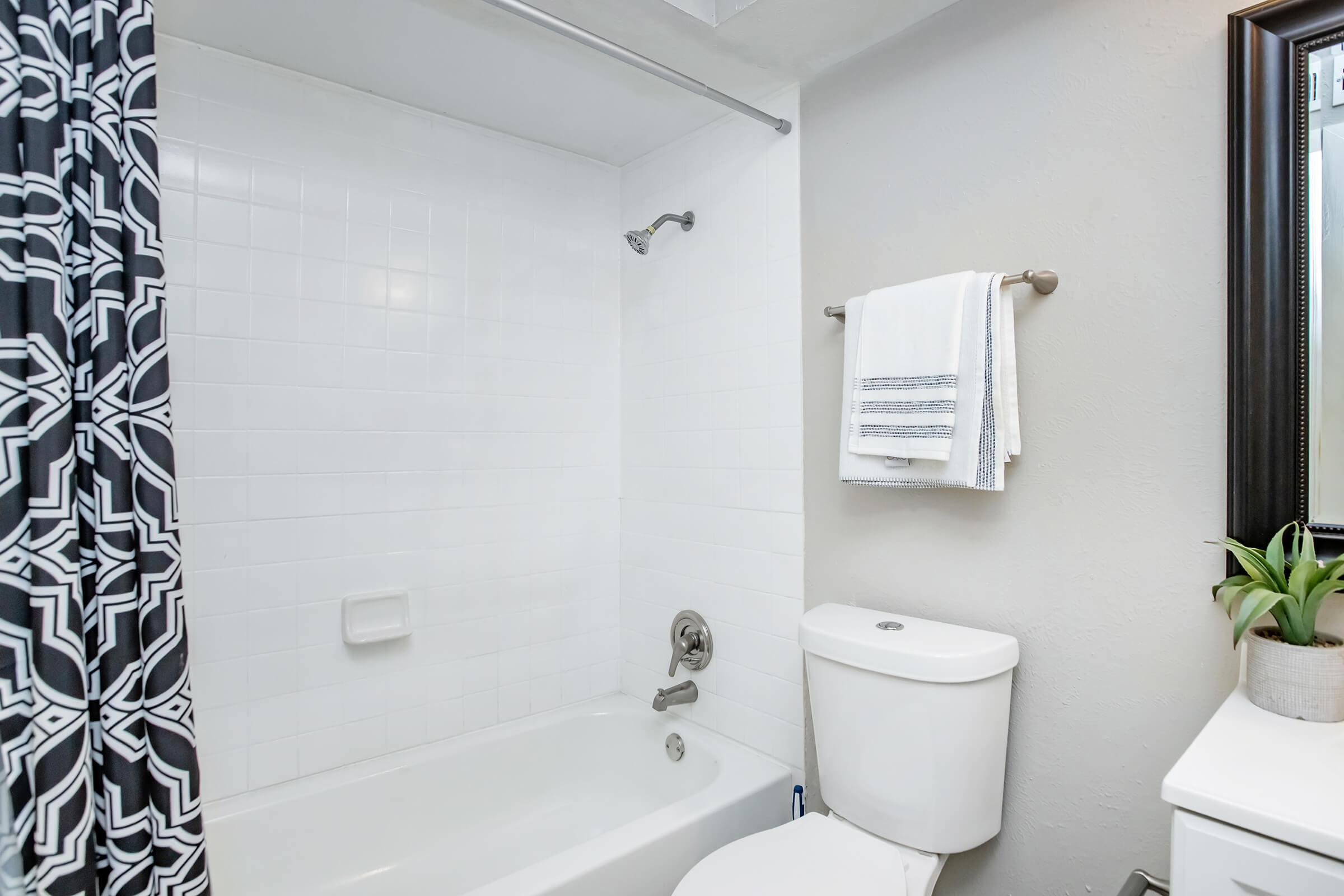 A modern bathroom featuring a bathtub with a showerhead, decorative black and white shower curtain, a white toilet, and two neatly folded towels on a towel rack. A small plant is displayed beside a mirror, contributing to the clean and simple aesthetic.