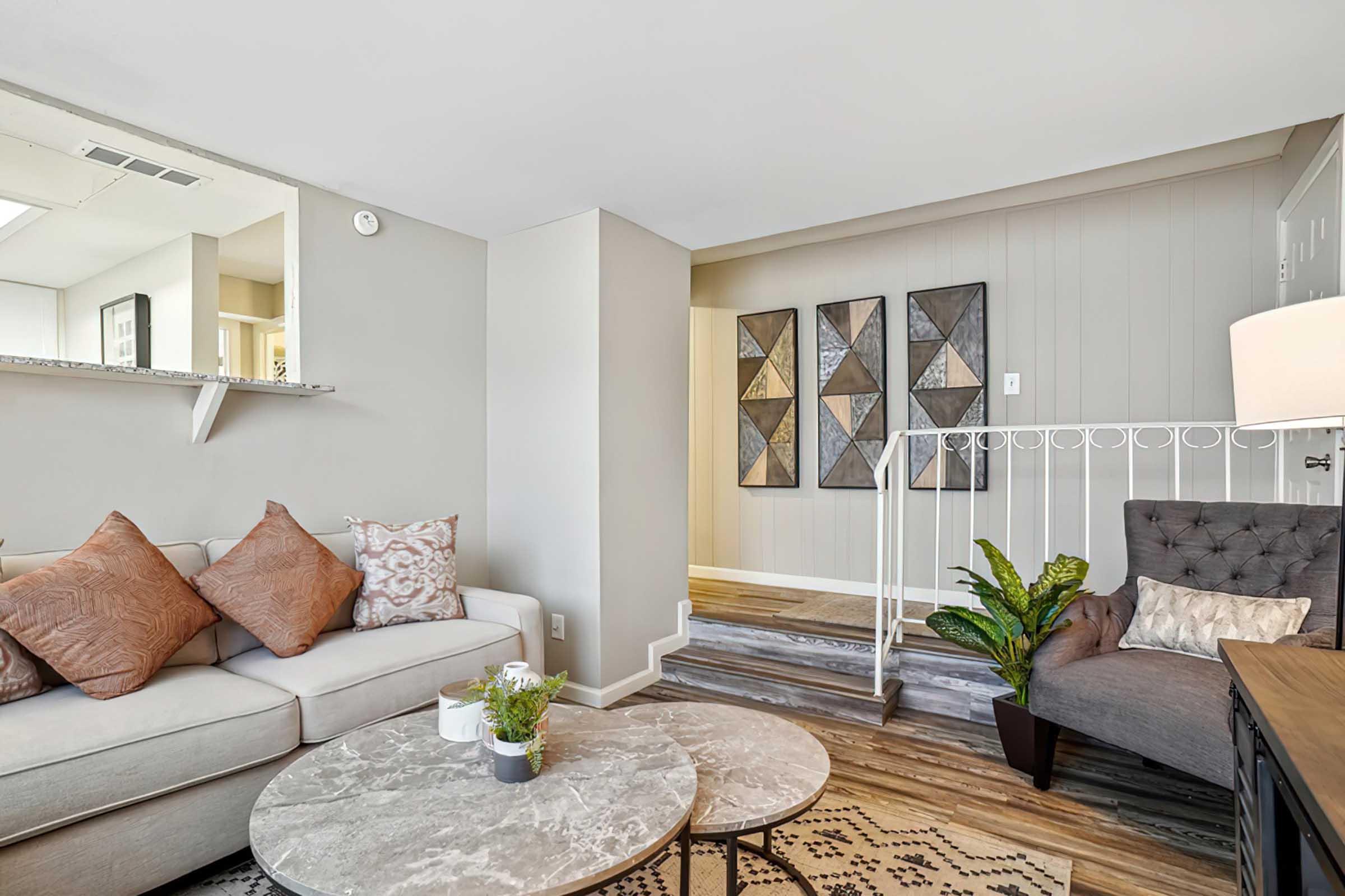 A cozy living room featuring a light-colored sofa adorned with decorative pillows, a round marble coffee table, and a plant. A stylish gray armchair is positioned nearby, with a small side table and lamp. The room is illuminated with natural light, and decorative wall art adds a modern touch. A staircase leads to another area.
