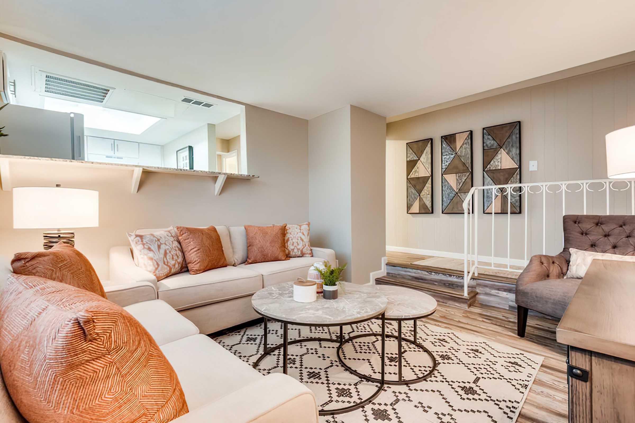 A modern living room featuring a light-colored sofa with orange accent pillows, a round coffee table in the center, and a patterned rug. There is a floor lamp nearby and decorative wall art composed of geometric shapes in the background. The space has warm, neutral tones and a cozy atmosphere.