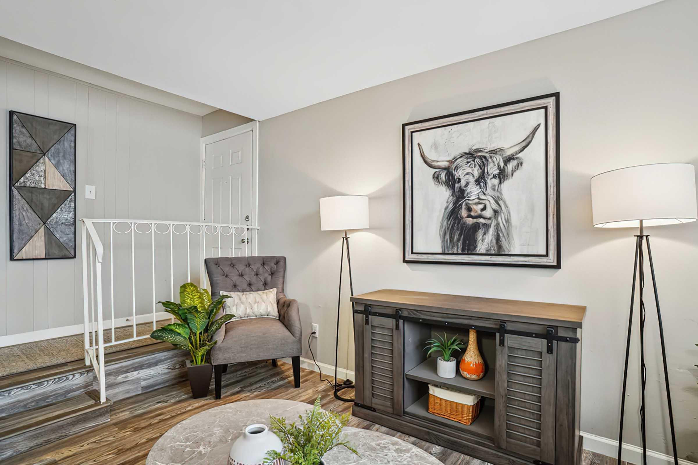 A cozy interior living space featuring a gray armchair, a decorative painting of a cow above a console table, plants, and a round marble coffee table. Soft lighting is provided by two table lamps, and a staircase leads to an upper level. The neutral color palette creates a warm atmosphere.