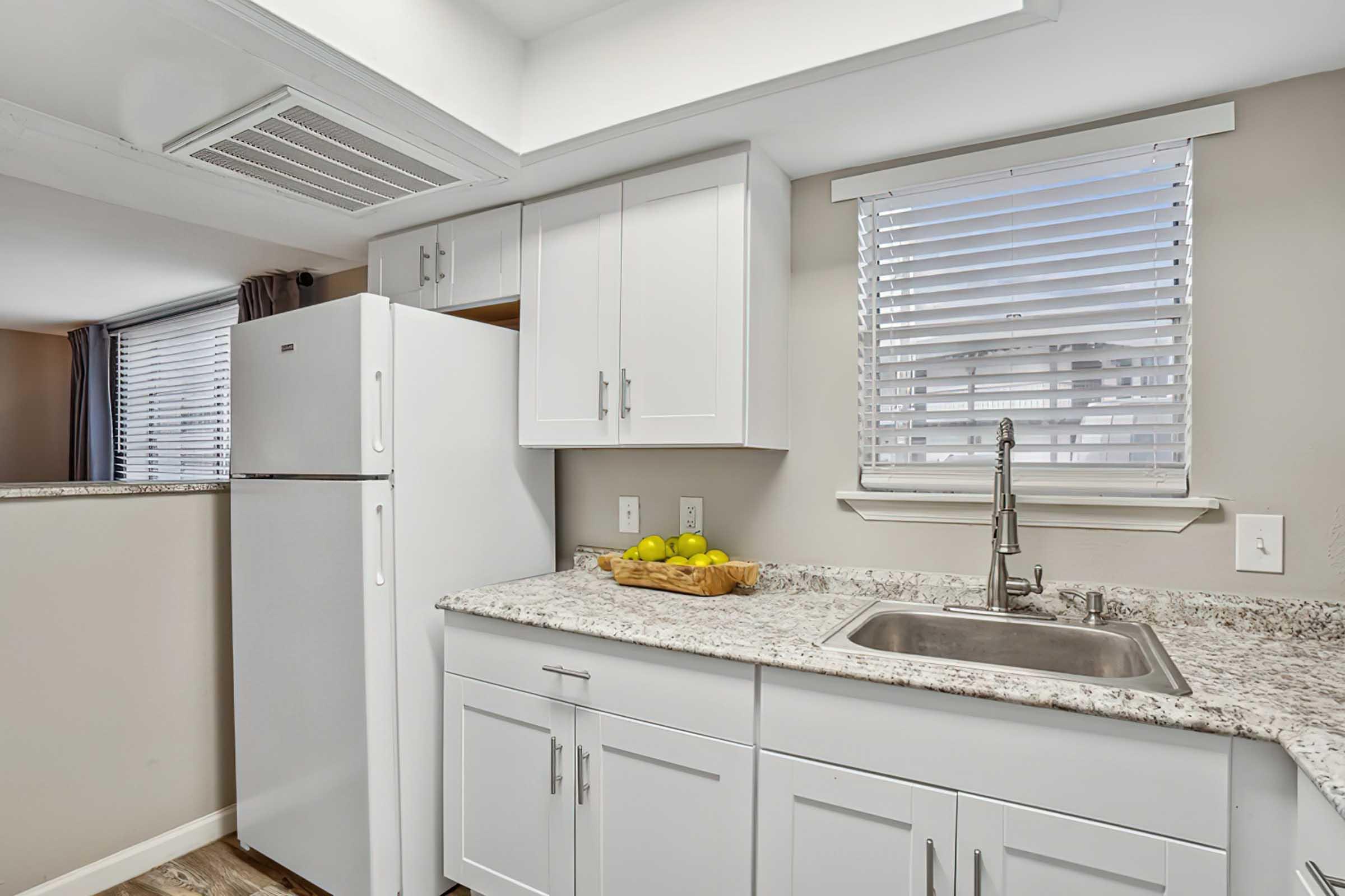 Bright, modern kitchen featuring a white refrigerator, a double sink with a sleek faucet, and a granite countertop. There is a wooden bowl with lemons on the counter, and the windows are adorned with white blinds, allowing natural light to brighten the space. The cabinets are white, enhancing the clean aesthetic.