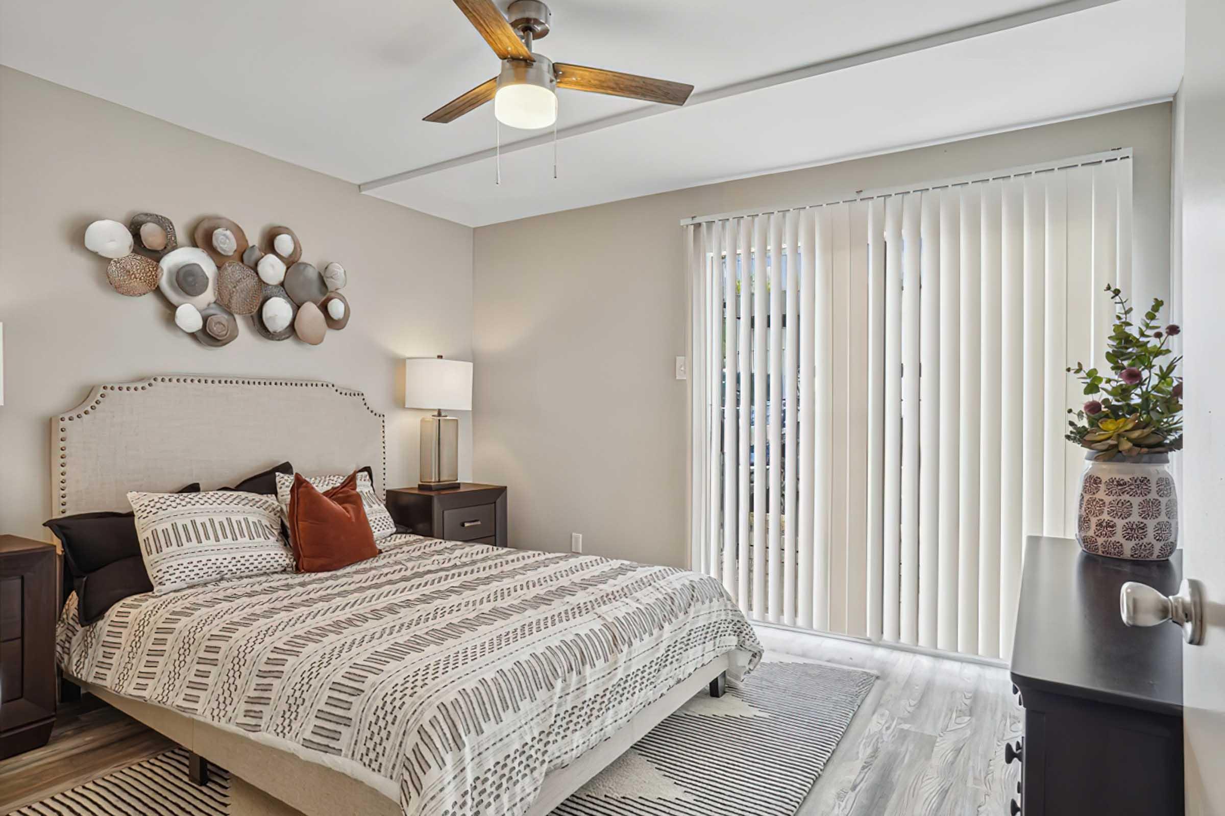A cozy bedroom featuring a queen-size bed with a textured duvet cover and decorative pillows. The room has a ceiling fan, bedside tables with lamps, and a large window covered with vertical blinds. Wall decor includes a circular arrangement of decorative plates. A plant is placed on a bedside table.