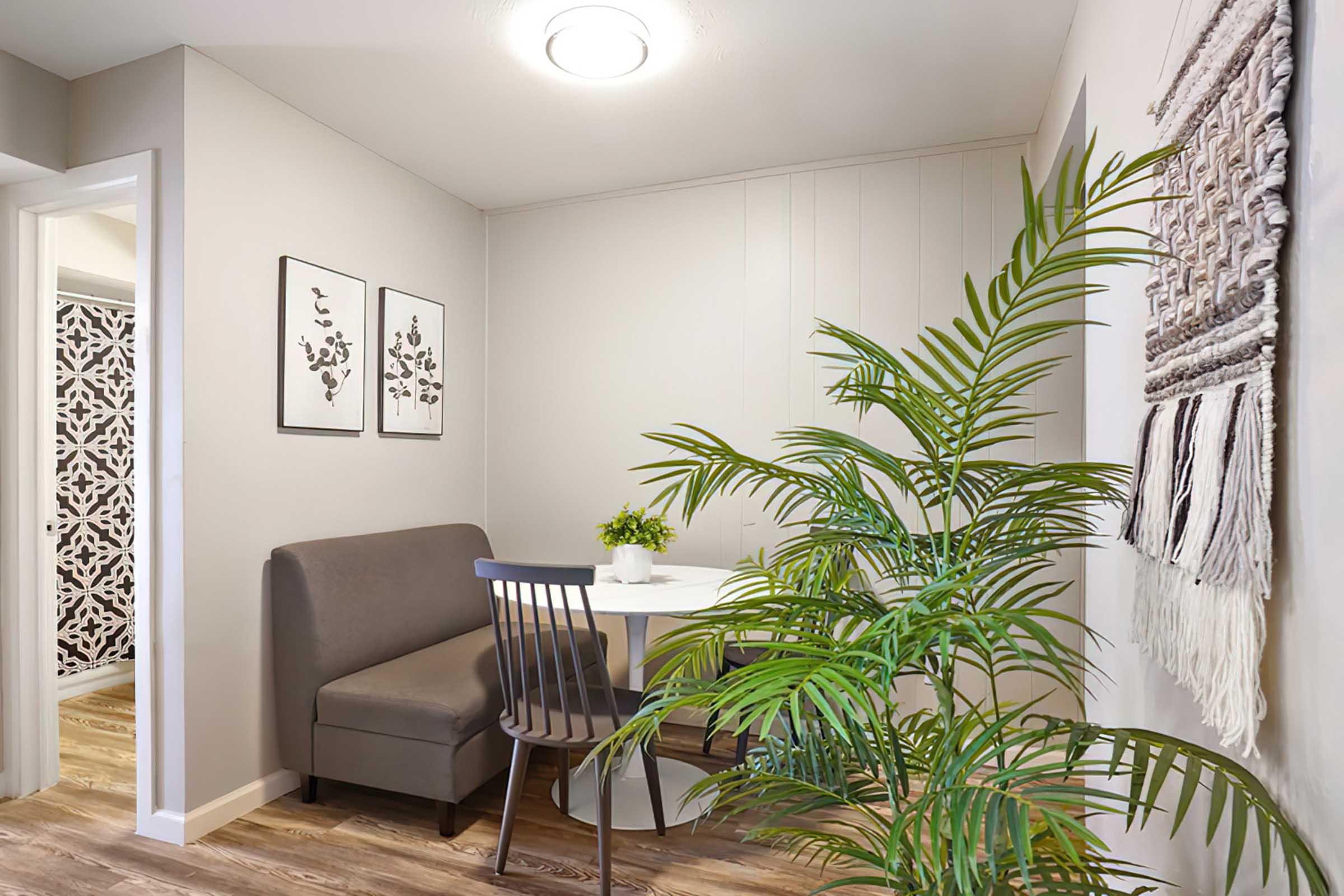 Cozy dining nook featuring a round table with a small potted plant, surrounded by a gray upholstered bench and black dining chair. Two framed floral prints adorn the wall, while a woven wall hanging adds texture. A leafy green plant brings a touch of nature to the space, set against light-colored walls and wood flooring.