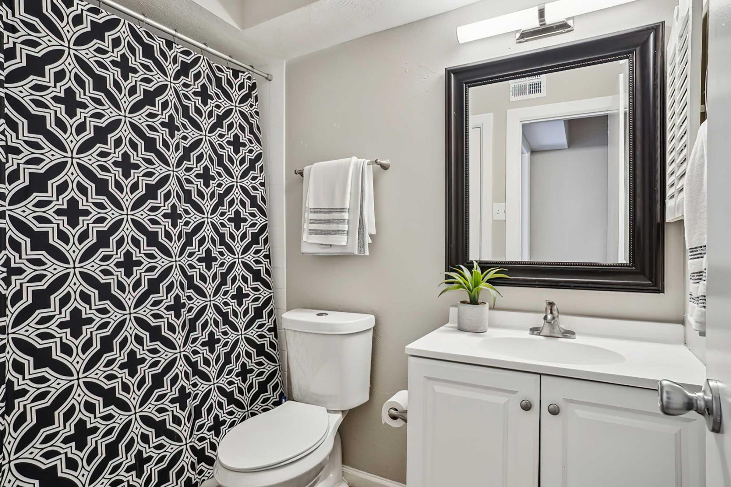 A modern bathroom featuring a black and white patterned shower curtain, a white sink with a mirror above it, and a toilet. The decor includes a small plant on the sink and a towel rack with neatly arranged towels. The walls are painted a light color, providing a bright and clean appearance.