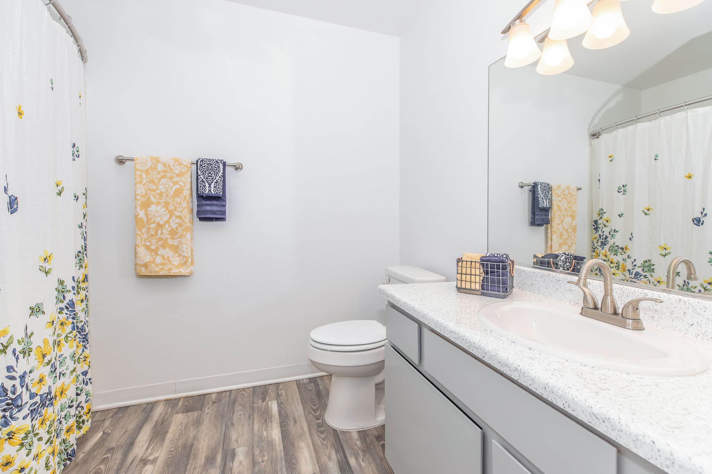 A clean and bright bathroom featuring a white sink with a granite countertop, a toilet, and a large mirror. The space includes a shower curtain with a floral pattern, two towels hanging on a rack, and a decorative basket on the counter, set against light walls and wood-like flooring.