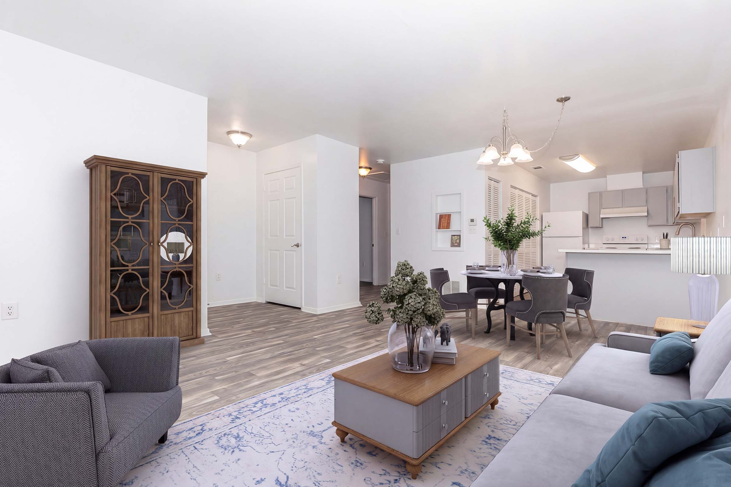 A modern, airy living space featuring a gray sofa and a wooden coffee table with decorative items. A dining area with a round table and gray chairs is visible in the background, along with a light-colored kitchen. The flooring is a light wood design, and there's a stylish cabinet against the wall.
