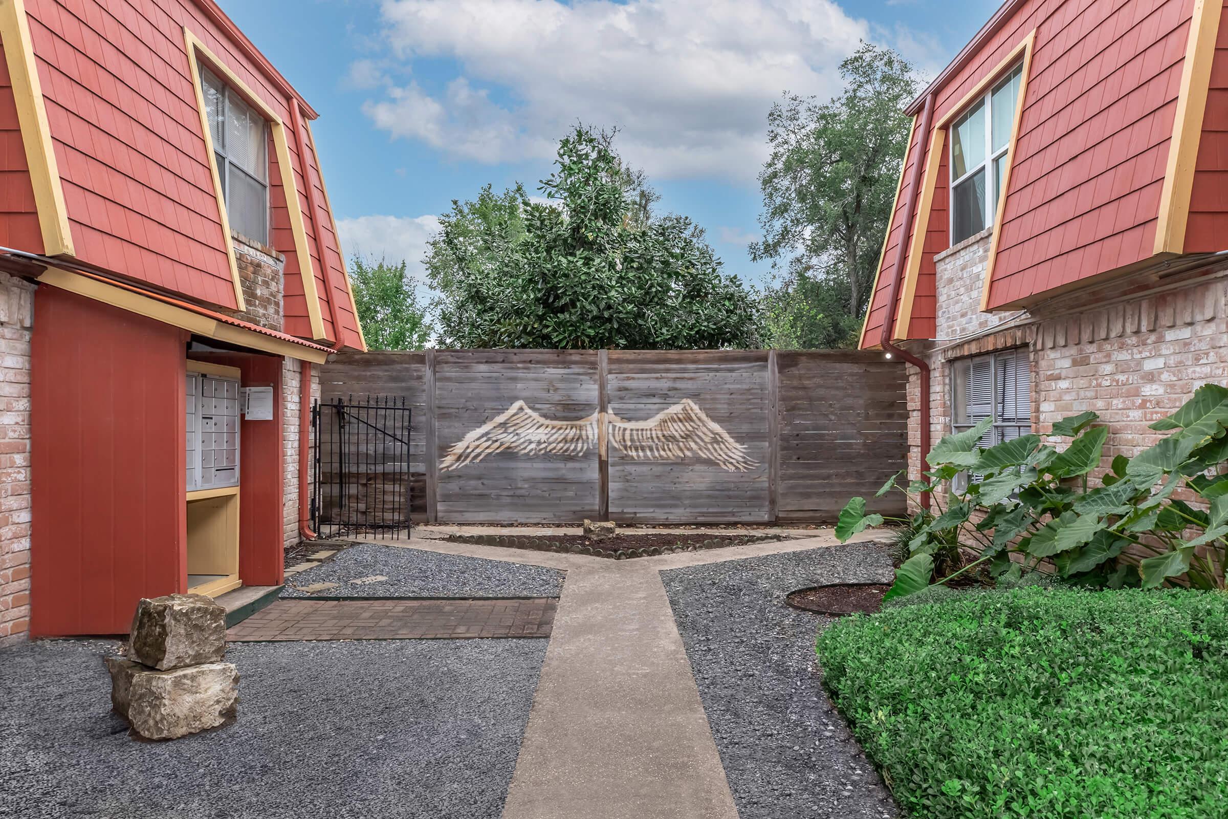 A courtyard between two buildings with red sloped roofs, featuring a wooden fence with a mural of angel wings. Lush greenery surrounds the pathways, and a stone sits near the entrance, creating an inviting atmosphere. The sky is partly cloudy, adding to the serene setting.