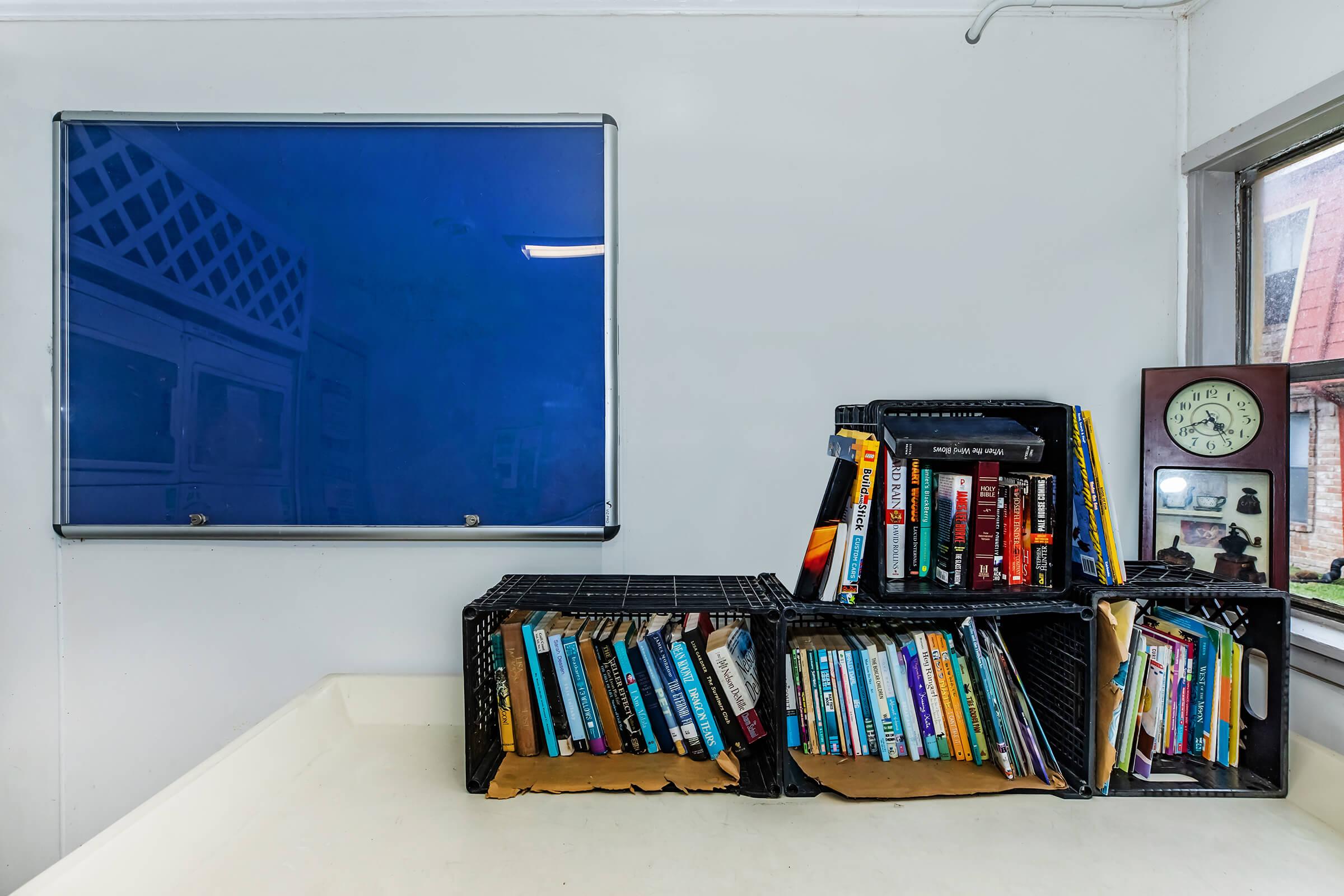 A cozy reading nook featuring stacked crates filled with books in various genres. A blue notice board hangs on the wall, and a clock is visible on a nearby shelf. The space has a bright, inviting atmosphere with soft natural light from a window.