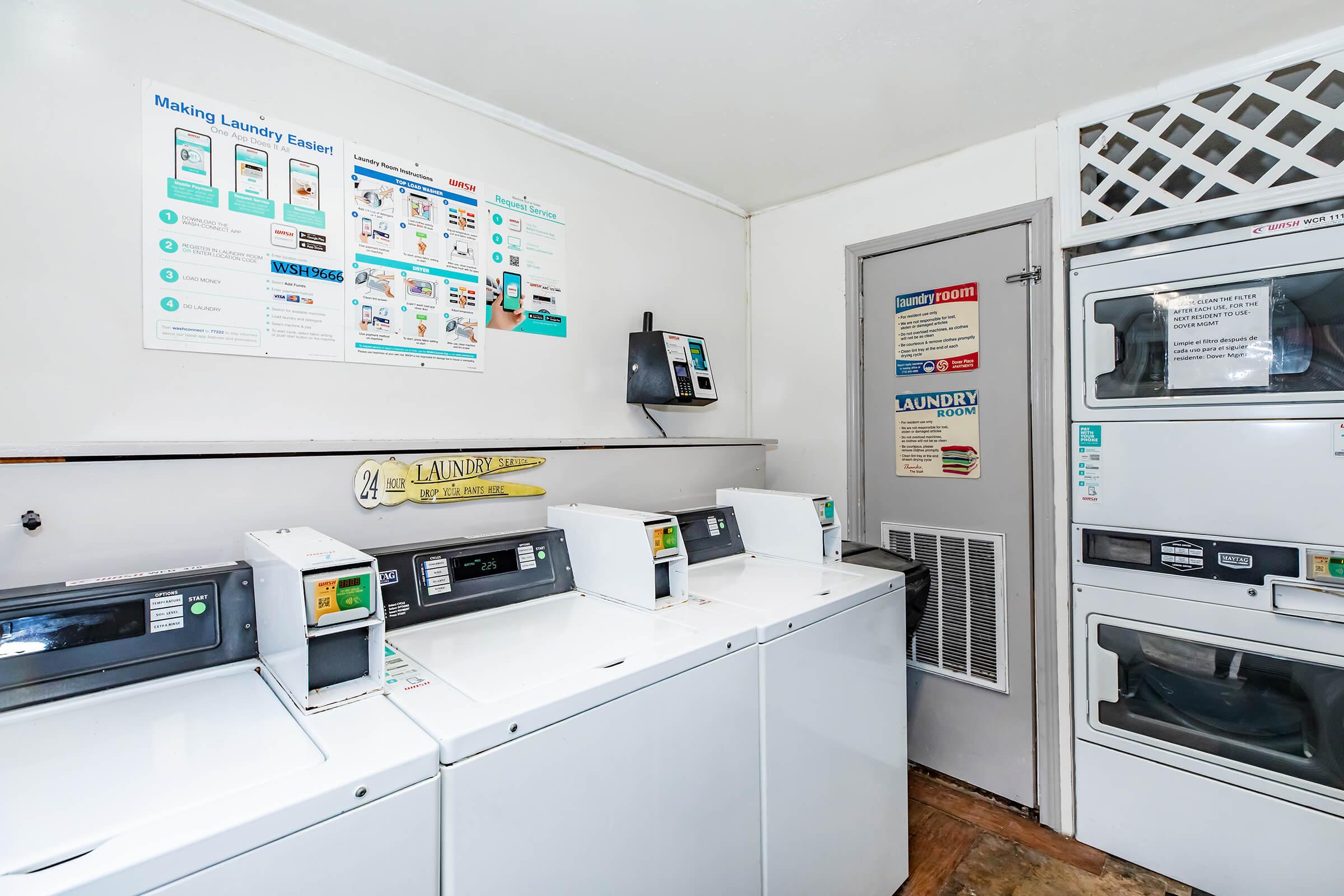A laundry room featuring several white washing machines and dryers. The walls display informational posters about laundry services and usage instructions. A vending machine is visible, and there is a door in the corner leading to another area. The floor has a simple, clean design.