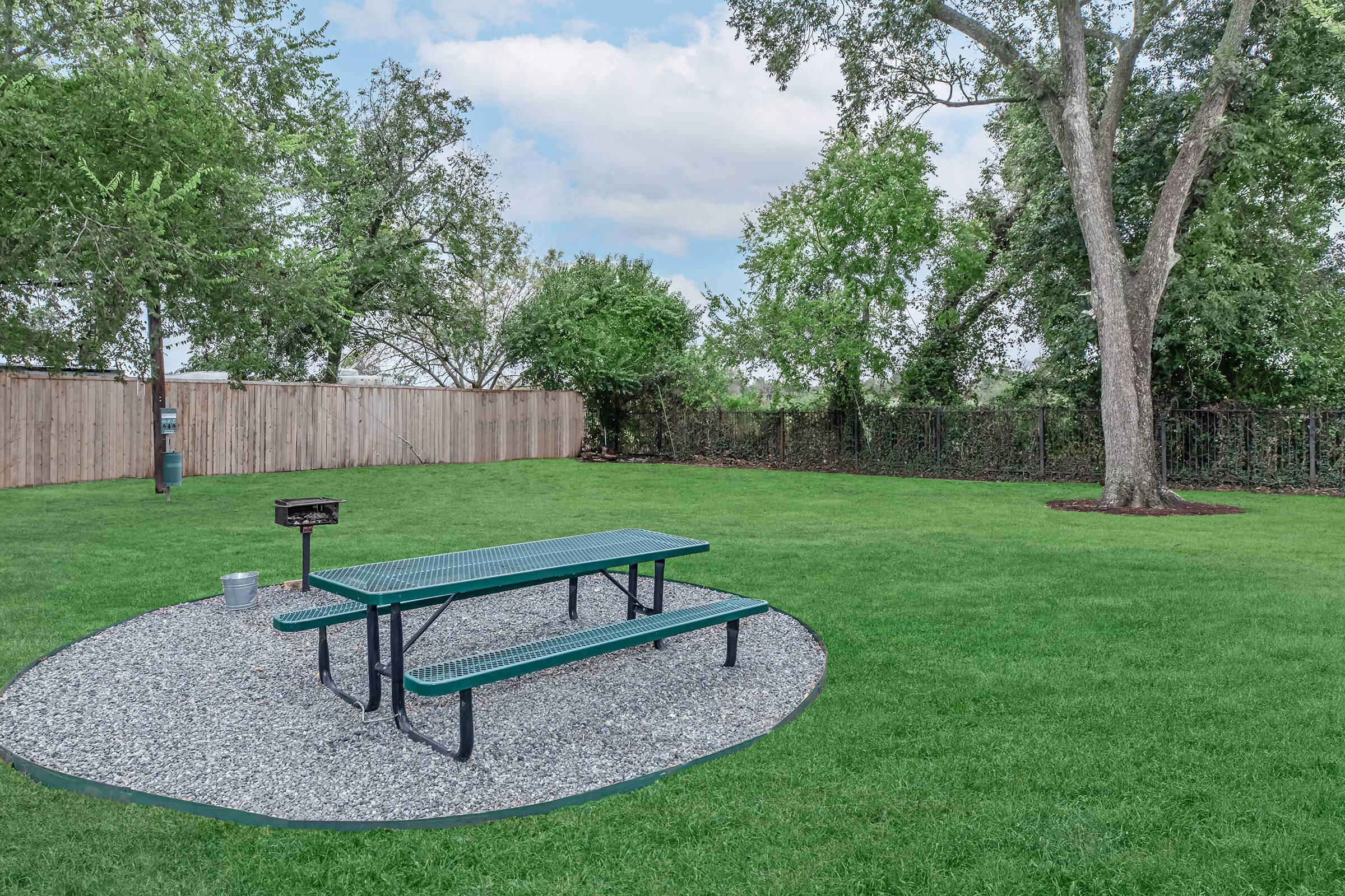 A landscaped backyard featuring a green picnic table on a gravel area, surrounded by lush grass and trees. A barbecue grill is visible nearby, with a wooden fence enclosing the space, and a clear blue sky partially covered by clouds above.
