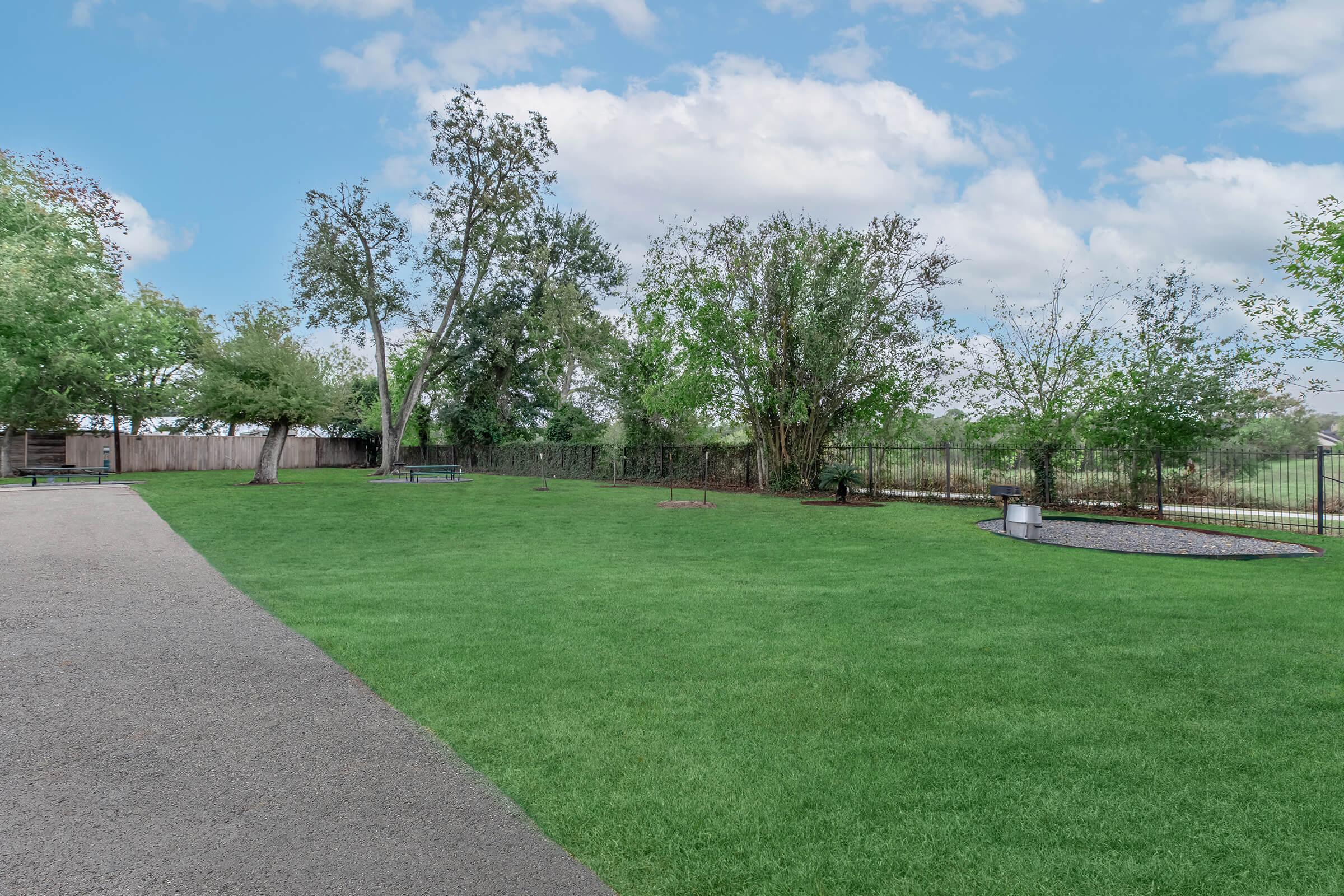 A spacious green lawn bordered by trees and shrubs, with a gravel path leading through the area. In the background, a fenced boundary and a small seating area made of gravel are visible. The sky is partly cloudy, enhancing the peaceful outdoor setting.