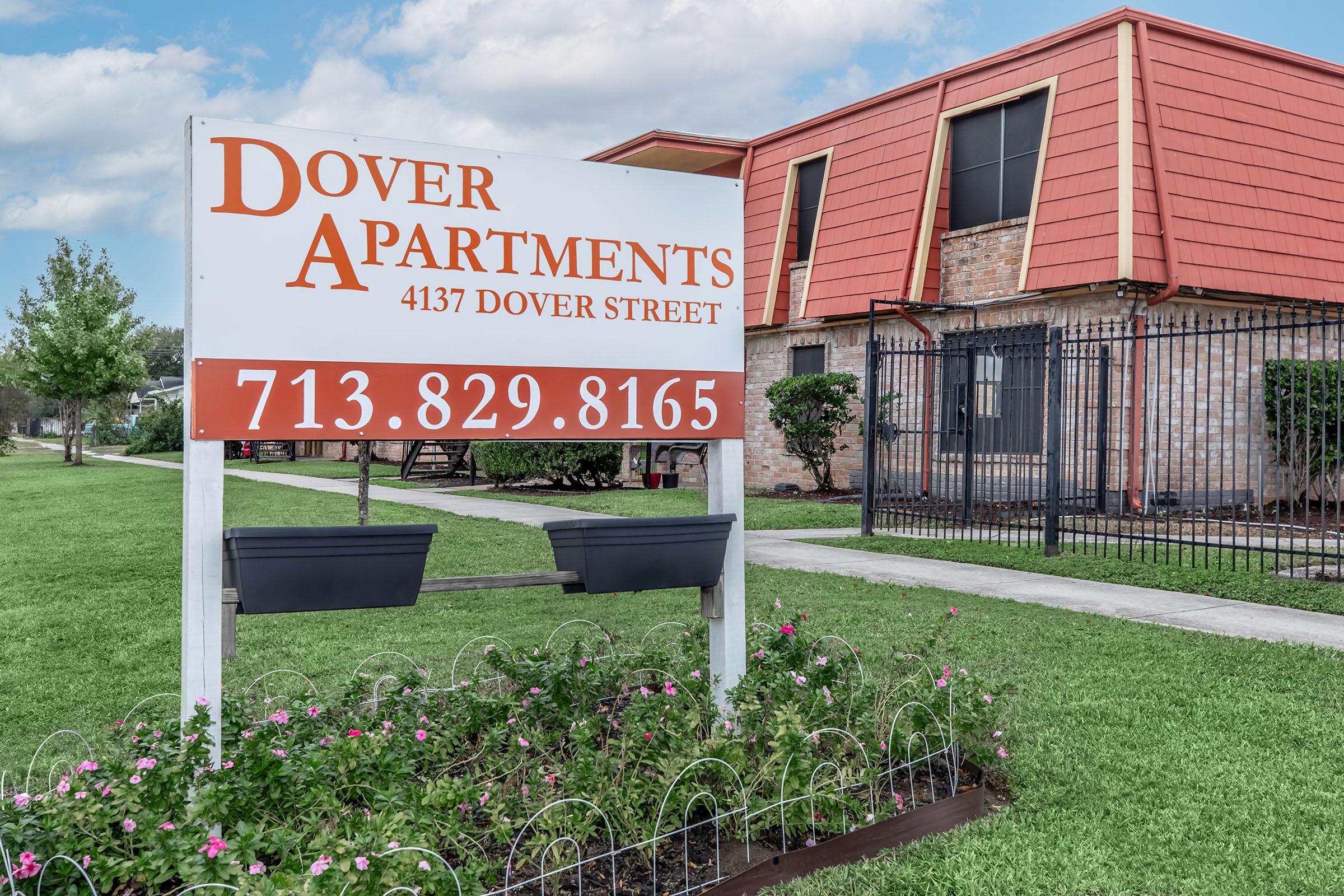 Sign for Dover Apartments located at 4137 Dover Street, featuring contact number 713-829-8165. The sign is prominently displayed in front of a brick building with an orange and brown roof, surrounded by a well-maintained lawn and flower beds.
