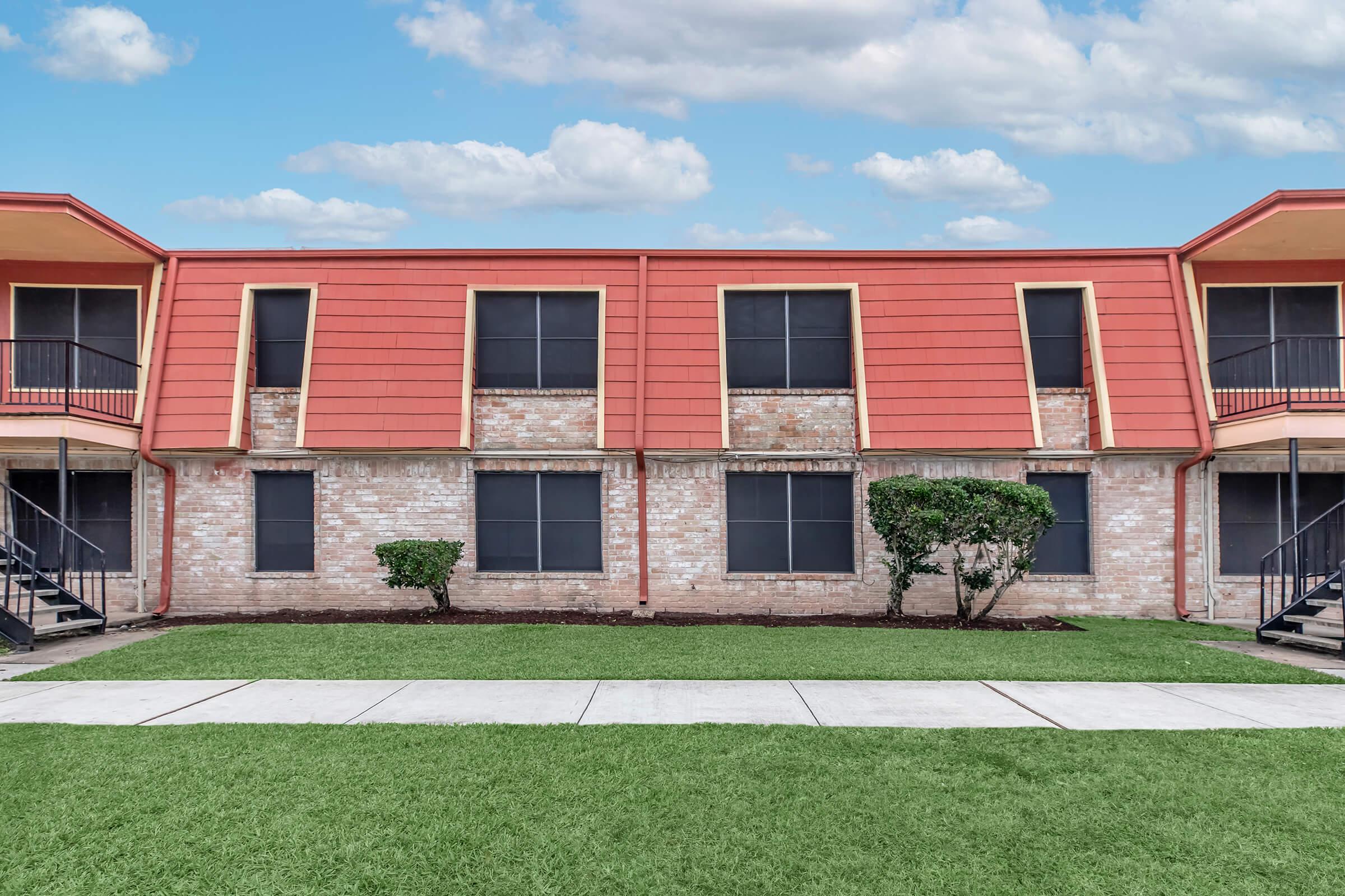 Exterior view of a two-story apartment building with a red sloped roof, featuring multiple windows and balconies. The building has a brick facade and is surrounded by well-maintained green grass. The sky is clear with scattered clouds, enhancing the bright atmosphere of the setting.