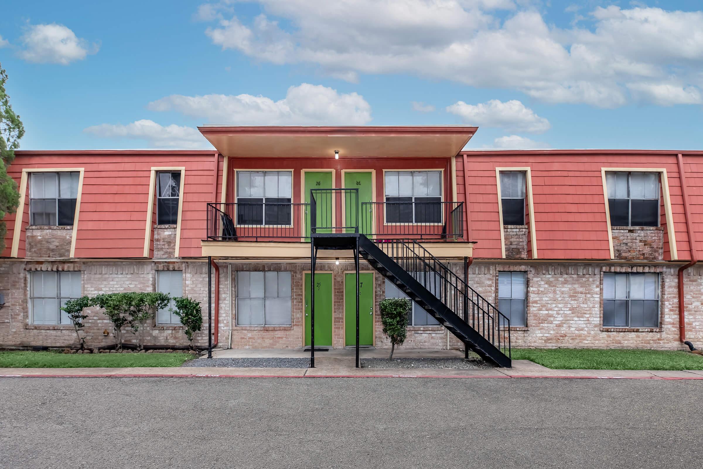 Two-story apartment building with a brick facade and bright orange and green exterior. A black staircase leads to the upper level, which has two doors. The ground level has windows on either side, and there are small shrubs along the walkway, under a mostly cloudy sky.