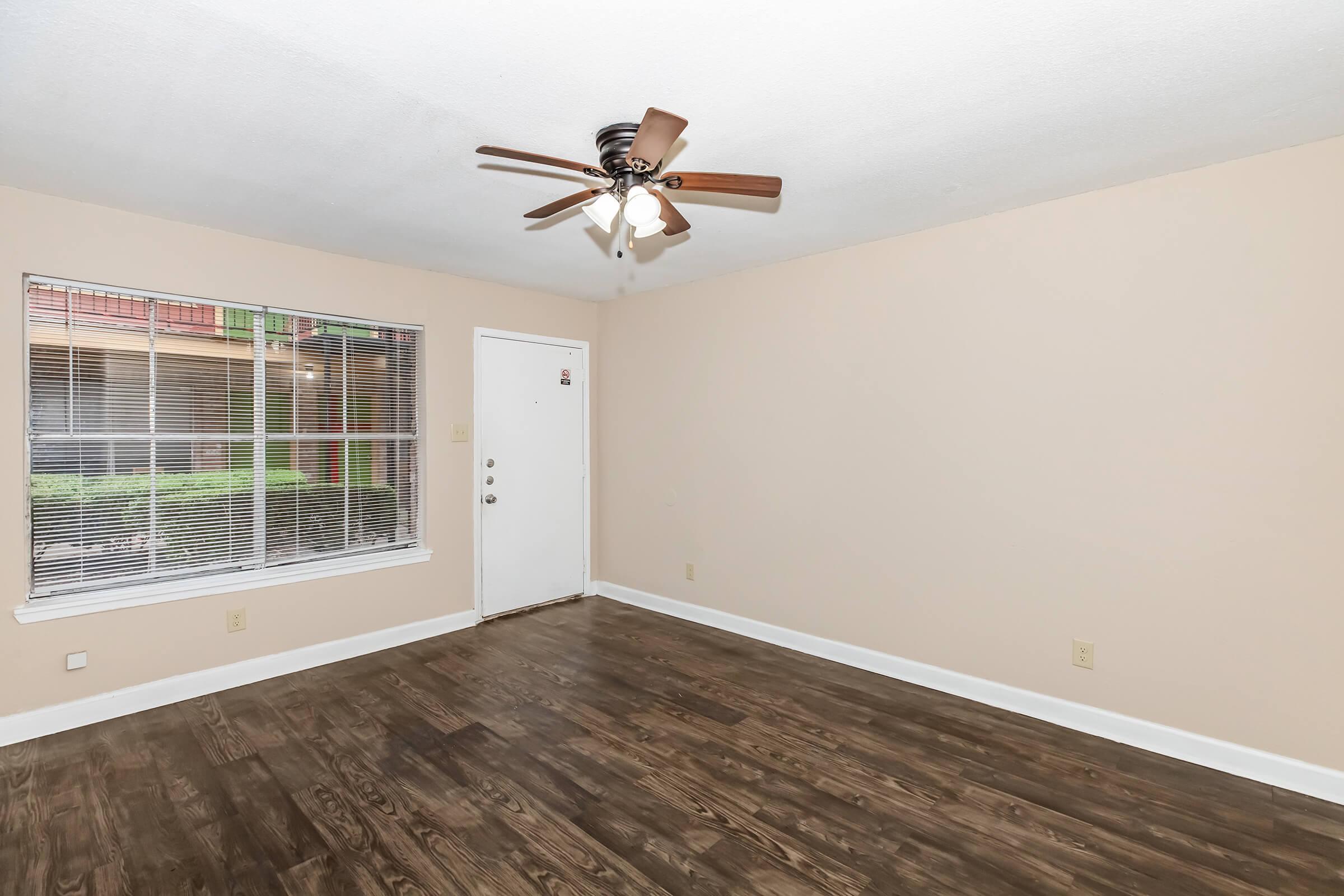 A spacious, unfurnished room featuring a light-colored wall and a wooden floor. A ceiling fan with wooden blades is installed, and a window provides natural light. The door leads outside, and the room appears ready for decoration or furniture placement.