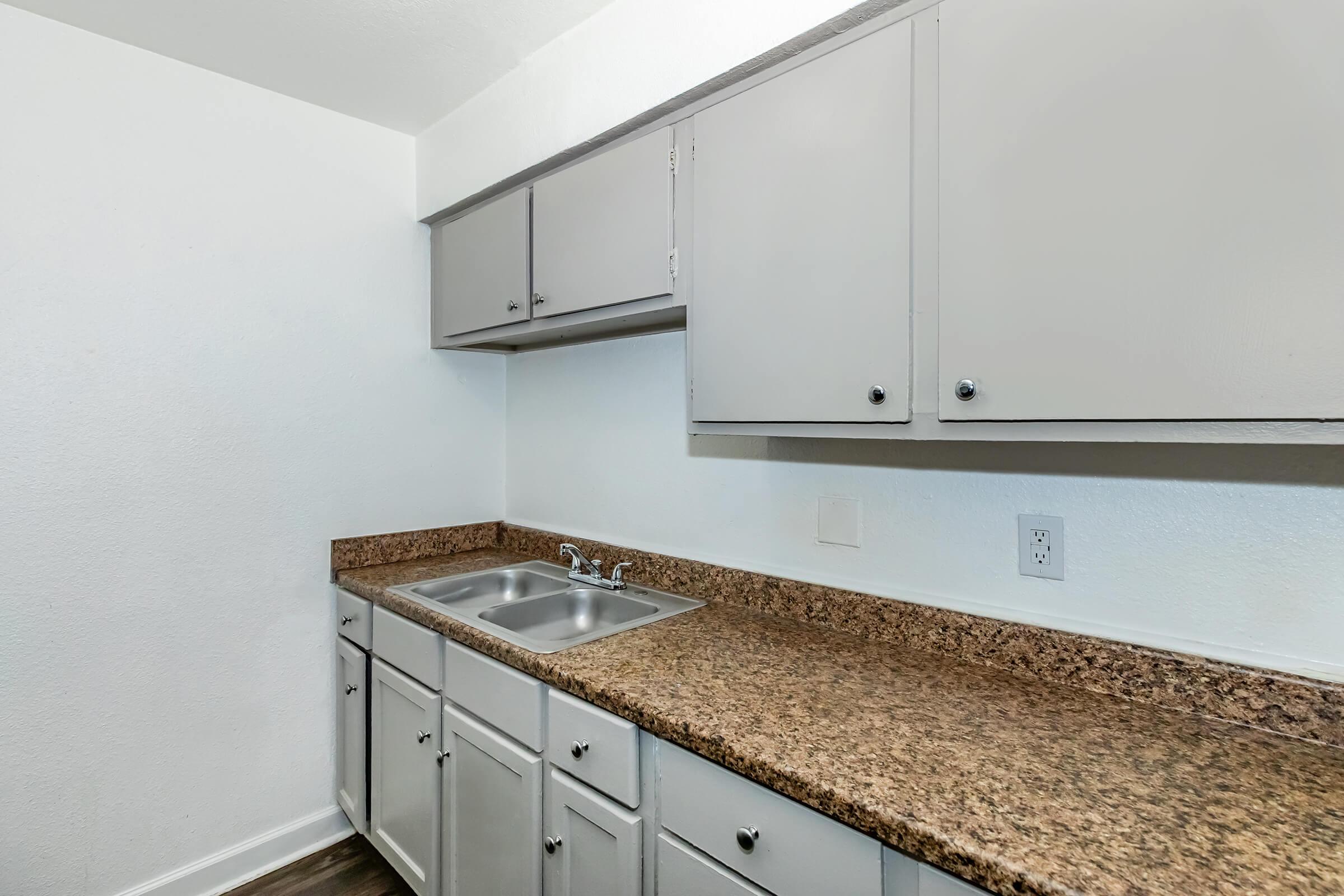 A clean kitchen space featuring light gray cabinets, a granite countertop, and a double sink. The walls are painted white, and the flooring appears to be a dark laminate. The overall design is simple and modern, suitable for a small apartment or home.
