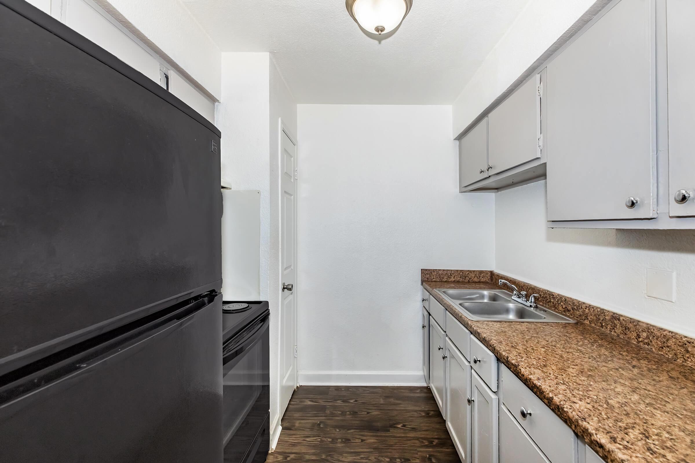 A modern kitchen featuring black appliances, including a refrigerator and stove, along with grey cabinets and a granite countertop. The space is well-lit with a ceiling light, and the flooring is dark wood. The kitchen has a sink and ample cabinet storage, creating a clean and functional area.