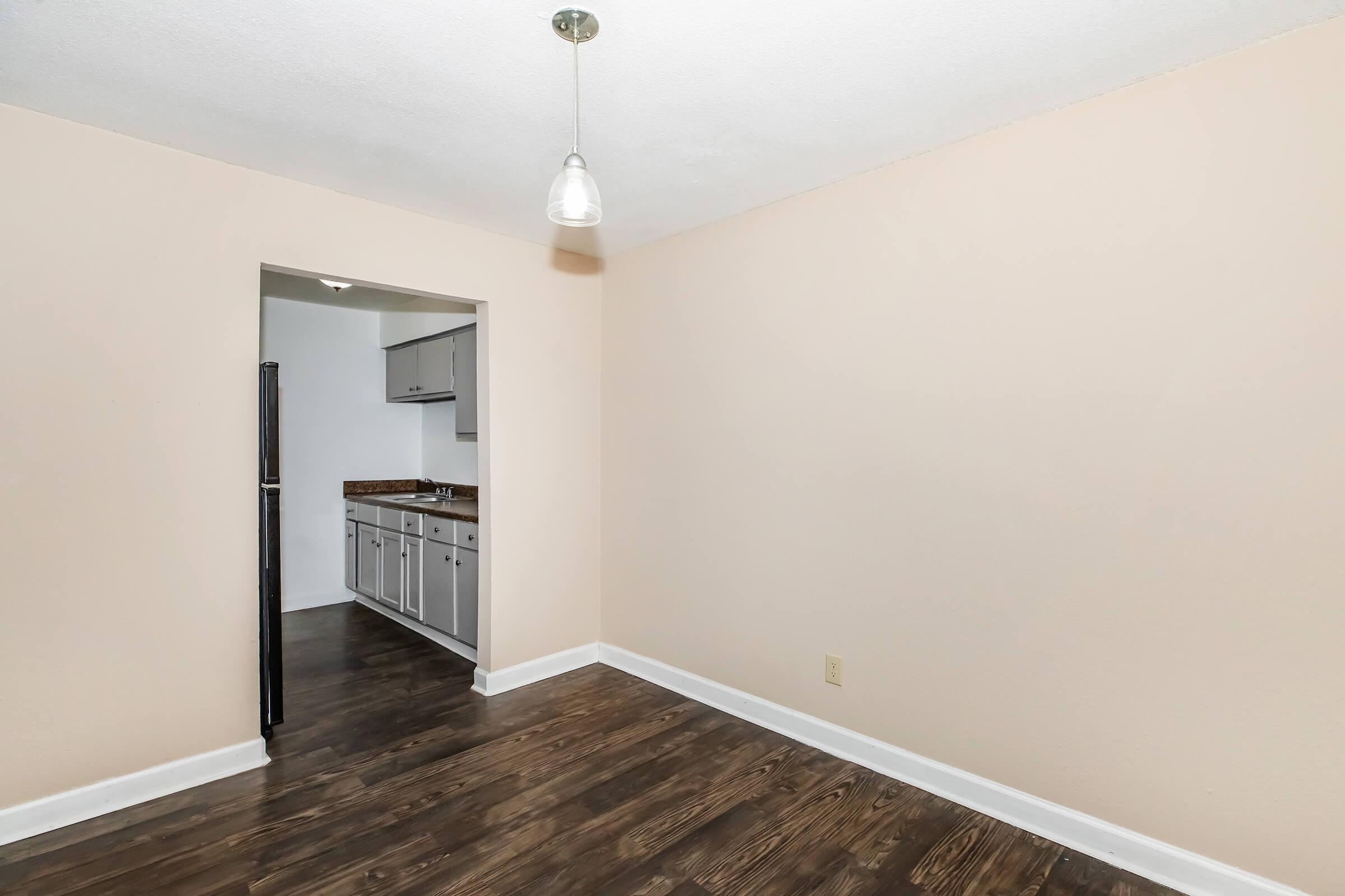 An empty room featuring light-colored walls and dark hardwood flooring. A doorway leads to a kitchen area with gray cabinets. A pendant light hangs from the ceiling, providing illumination. The space appears clean and ready for furnishings.