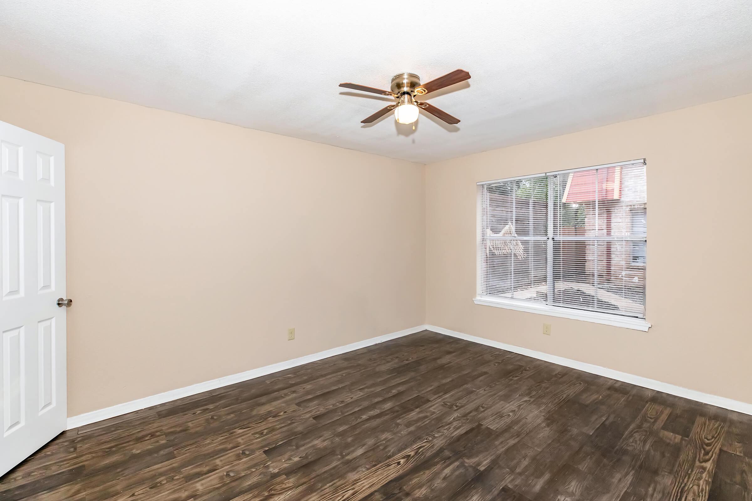 A spacious, empty room with light brown walls and a window featuring blinds. The room has a ceiling fan with a light fixture and a wooden floor. A white door is visible on the left side, and the overall atmosphere is bright and clean, ideal for use as a living space or bedroom.