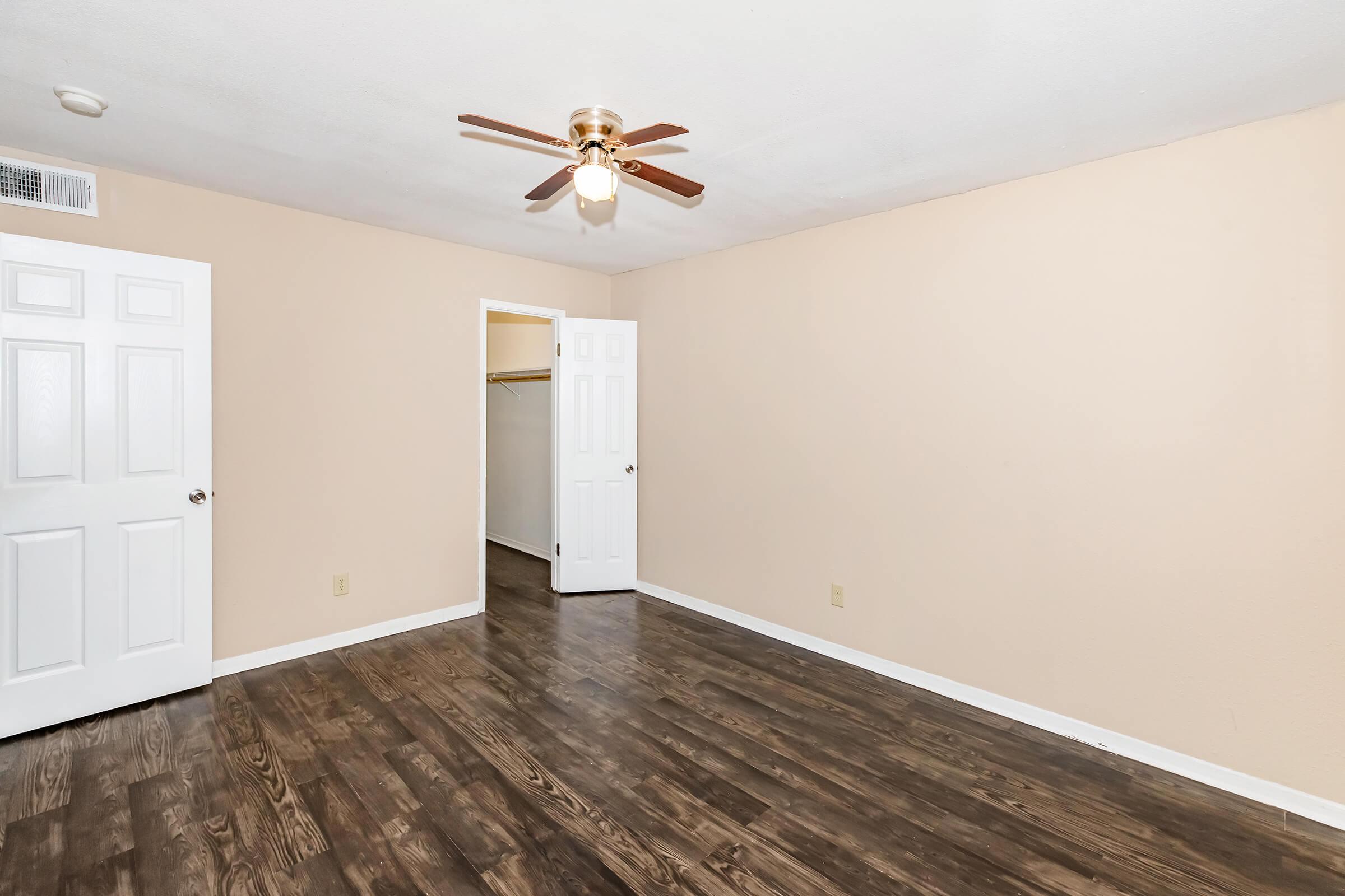Empty room featuring light beige walls, a wooden floor, and a ceiling fan. There are two white doors: one leading to a closet and the other to another room. The space is bright and spacious, ideal for various furnishings and decor.