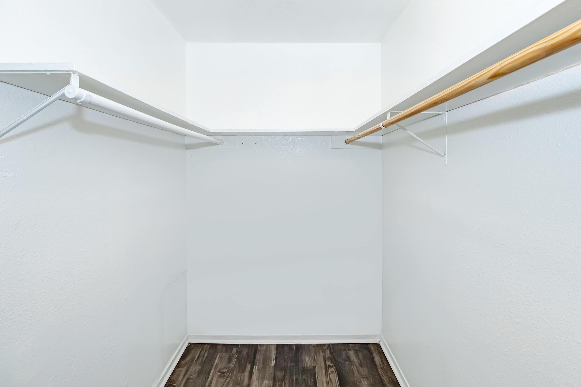 Empty closet with white walls and a dark wood floor. There are two hanging rods: one white and one wooden. The space is minimalist and uncluttered, creating a clean and open feel.