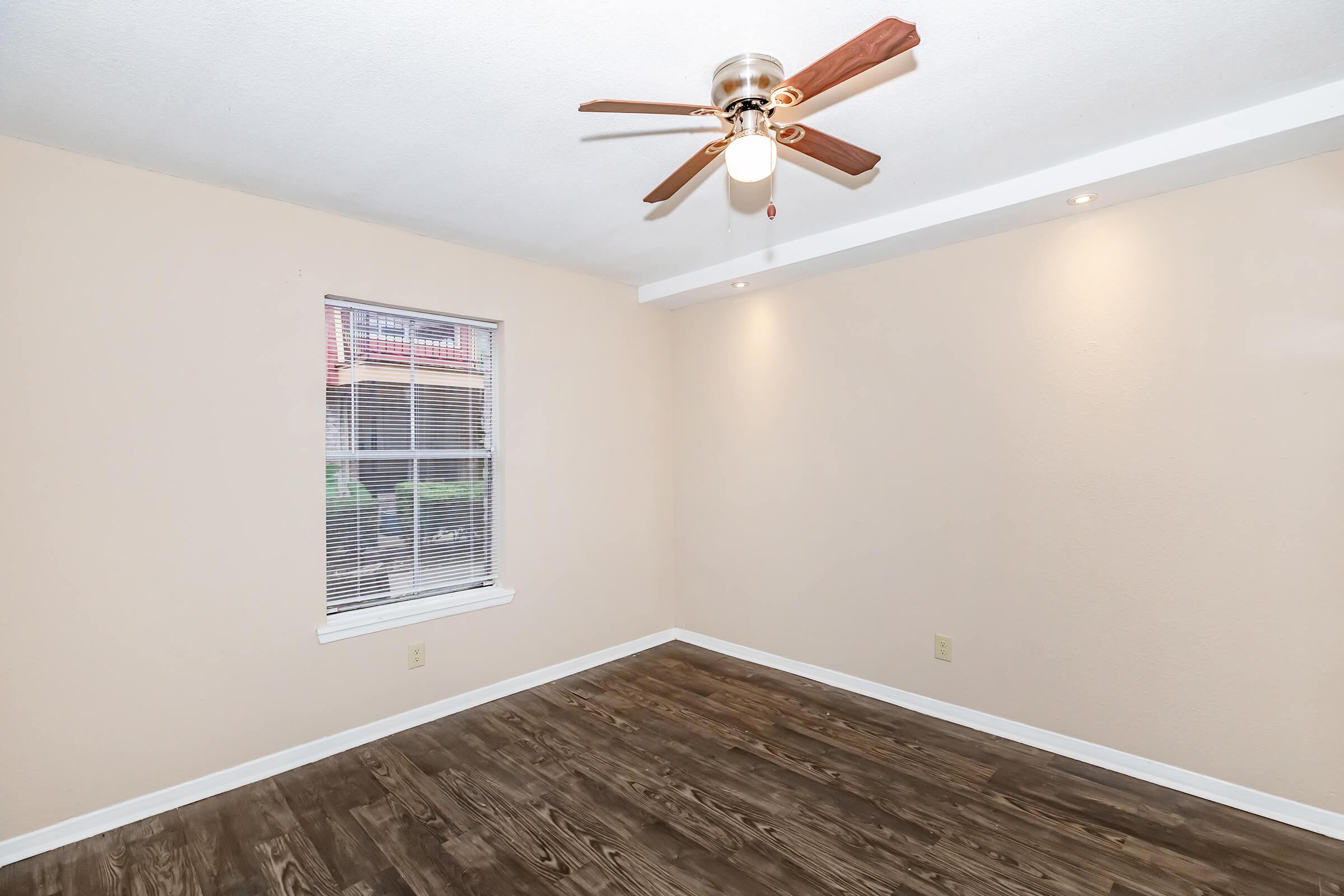 A small, empty room with light beige walls and wooden flooring. It features a ceiling fan with wooden blades and a light. There is a single window with blinds allowing natural light to enter. The room is well-lit and has minimal decor, providing a neutral space ready for furnishing.
